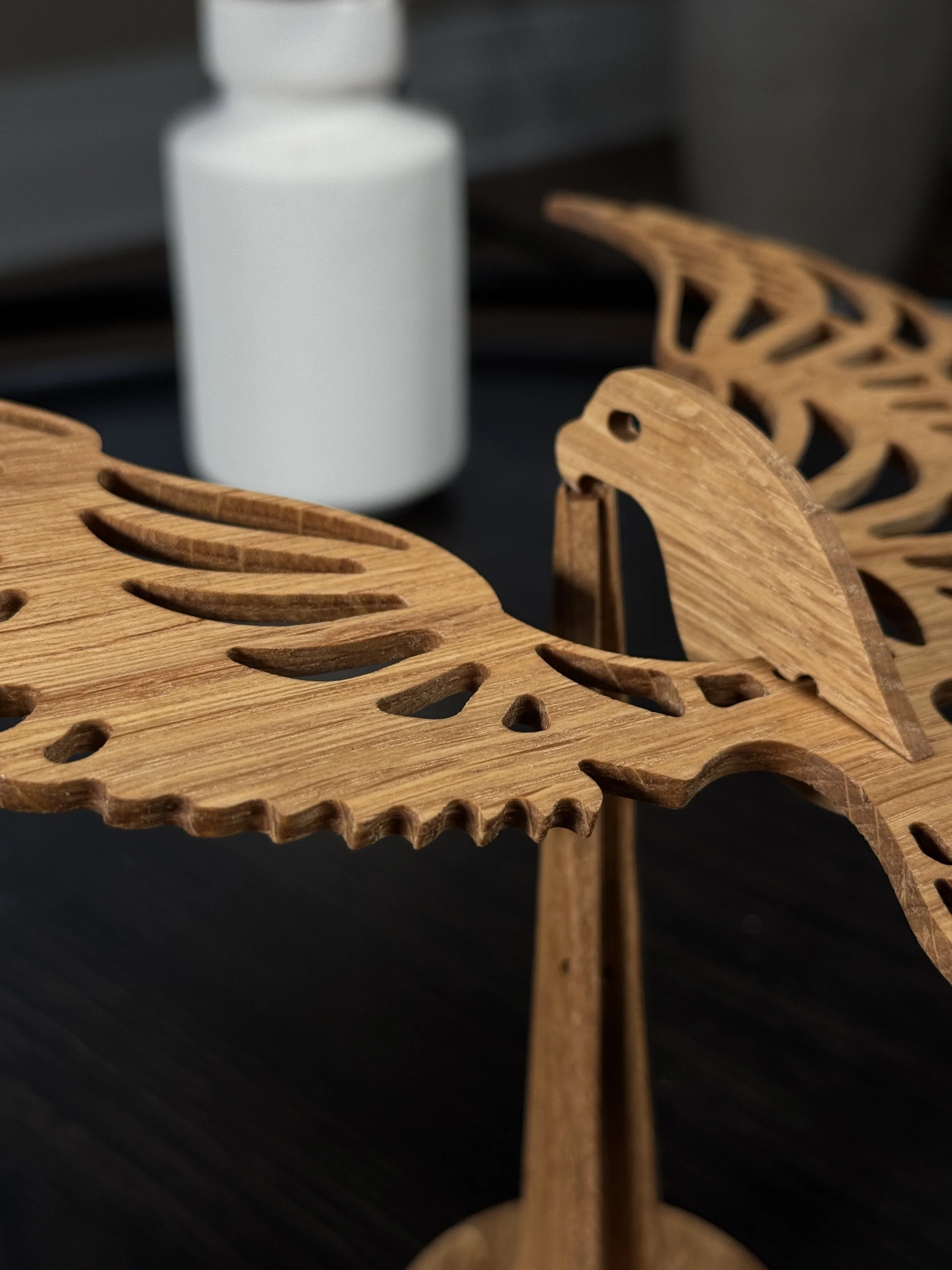 Close-up of a wooden decorative piece with leaf and bird cutouts, placed on a dark surface with a white vase in the background.