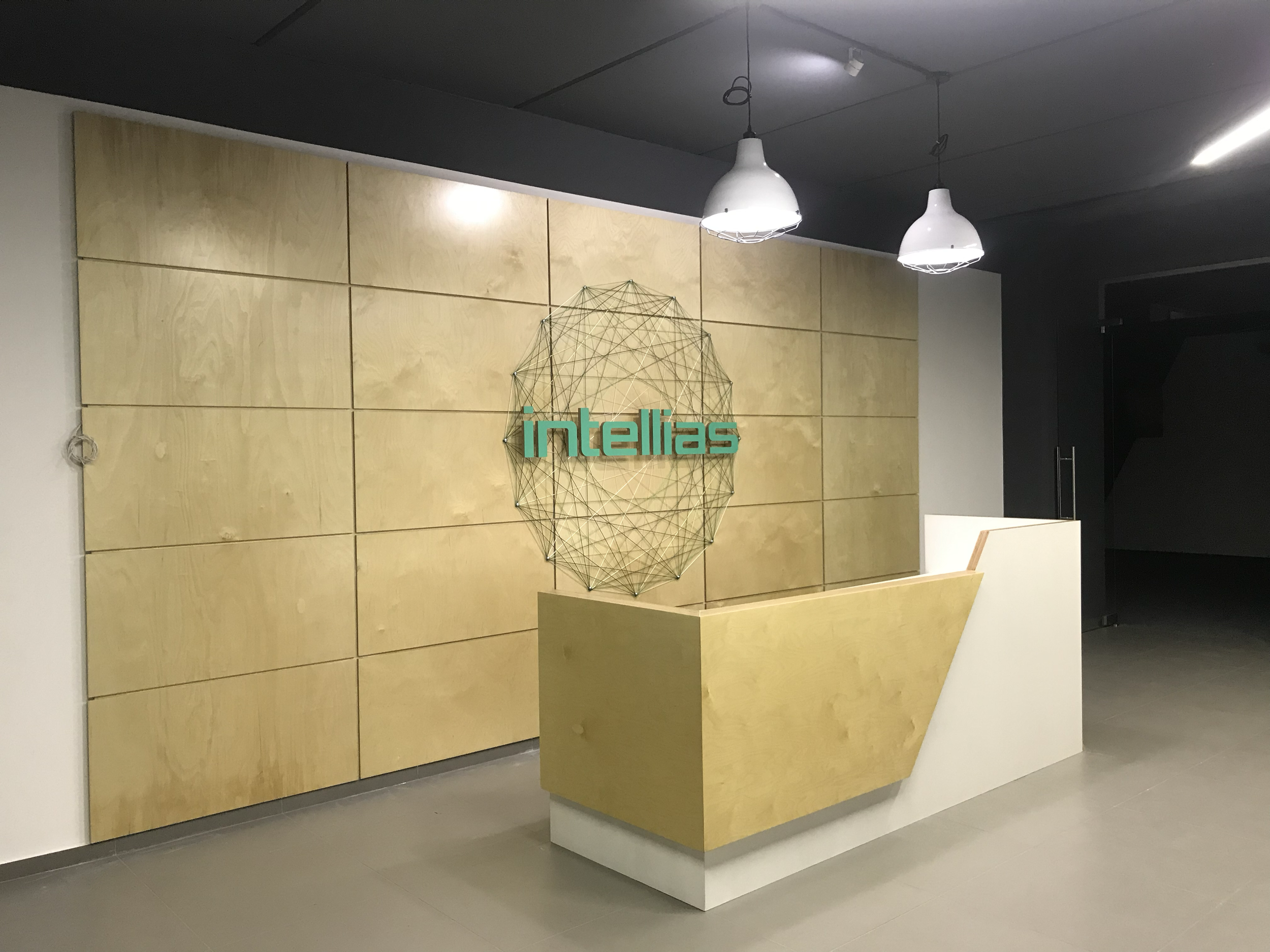 Office reception area with a wall display that has the word 'Intellias' and a geometric wireframe design, a modern angular reception desk, and two hanging white pendant lights.
