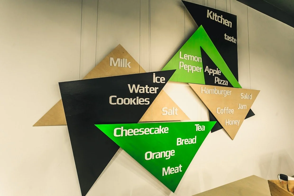 Decorative wall with colorful geometric shapes displaying various food and drink items, including milk, ice water, cookies, salt, lemonade, pepper, apple, pizza, hamburger, salad, jam, coffee, honey, cheesecake, tea, bread, orange, and meat, amidst a