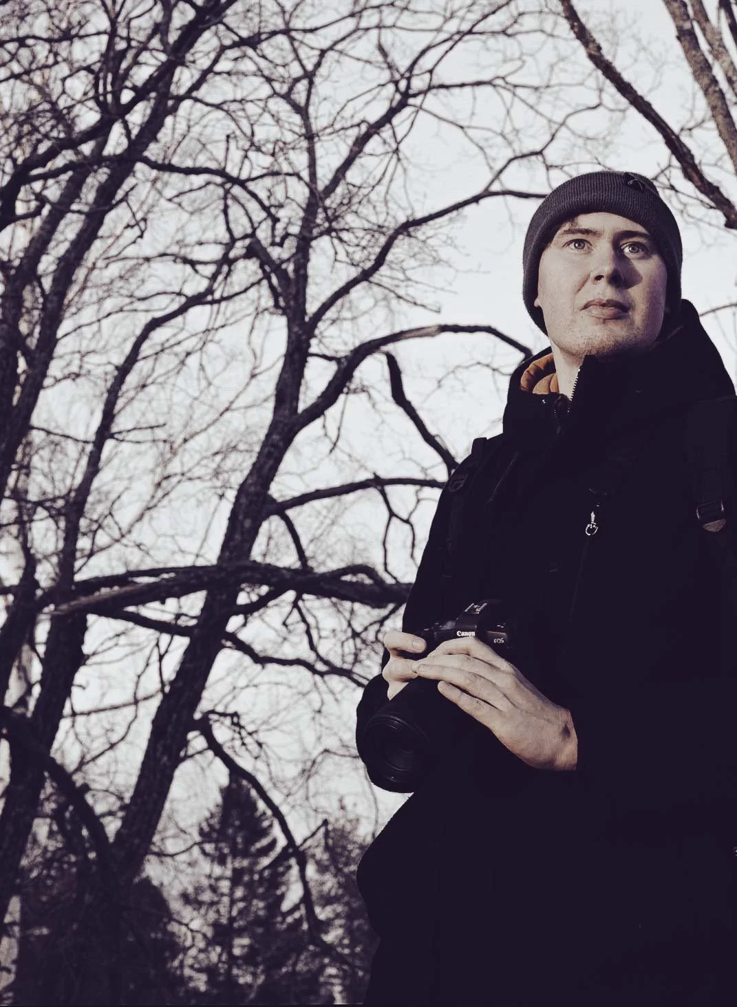 A man wearing a black jacket and gray beanie holding a camera during the daytime, standing outdoors in front of leafless trees.
