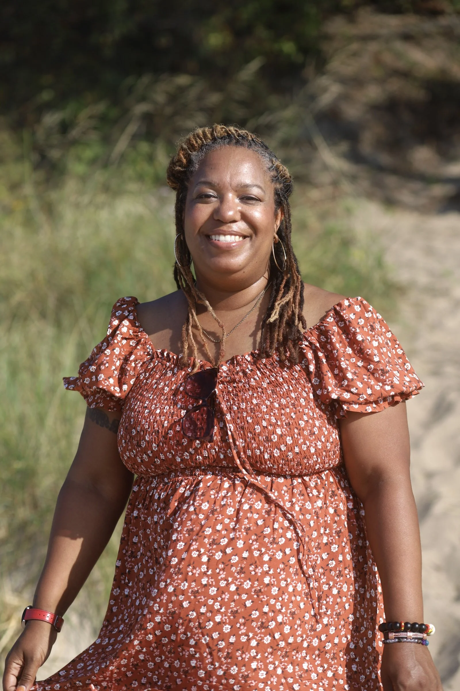 A smiling woman with dreadlocks wearing a red floral dress and sunglasses hanging from her chest, standing outdoors on a sunny day.