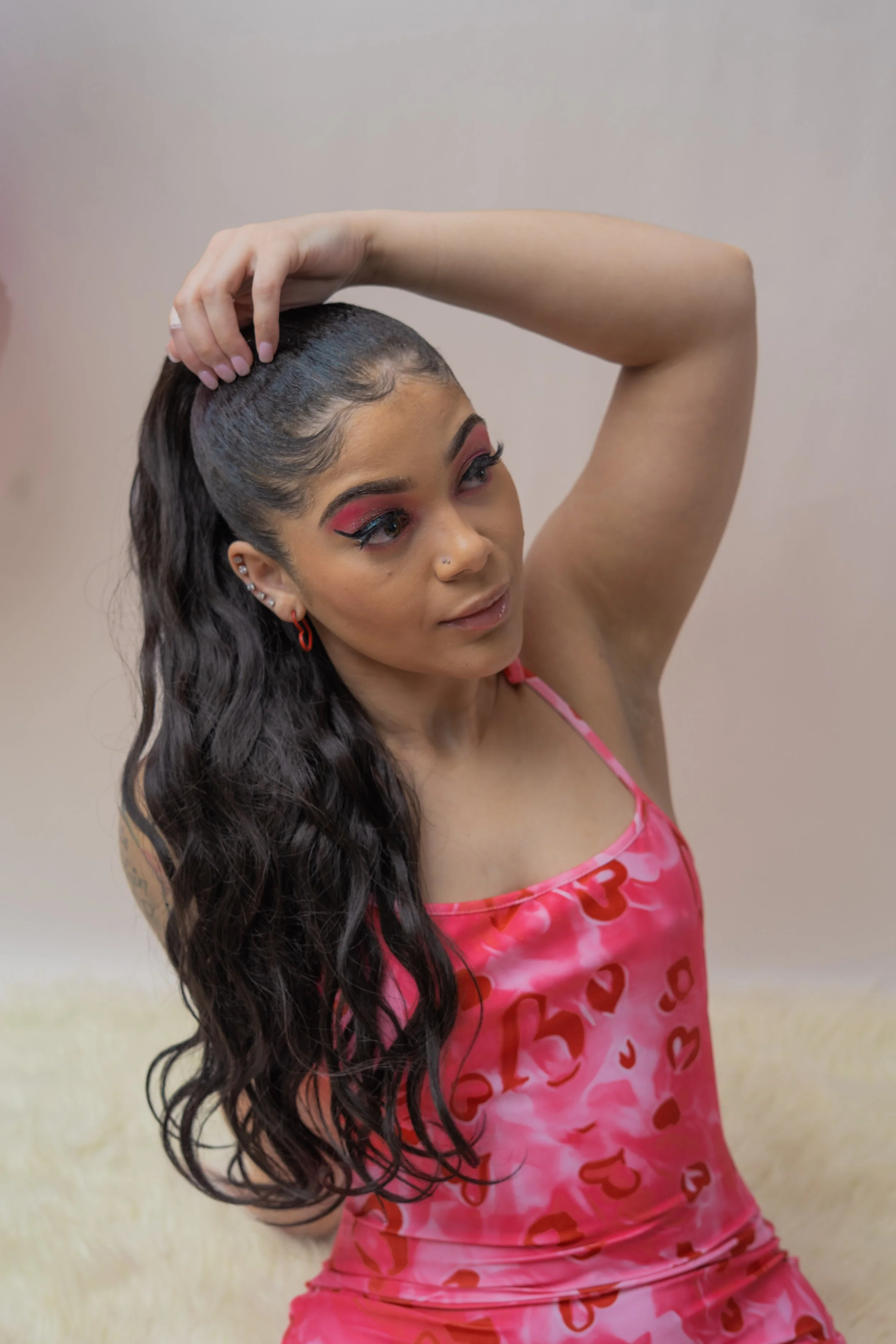 A young woman with long, wavy dark hair, wearing pink makeup and a pink dress with heart patterns, posing with her right arm raised and her hand on her head.