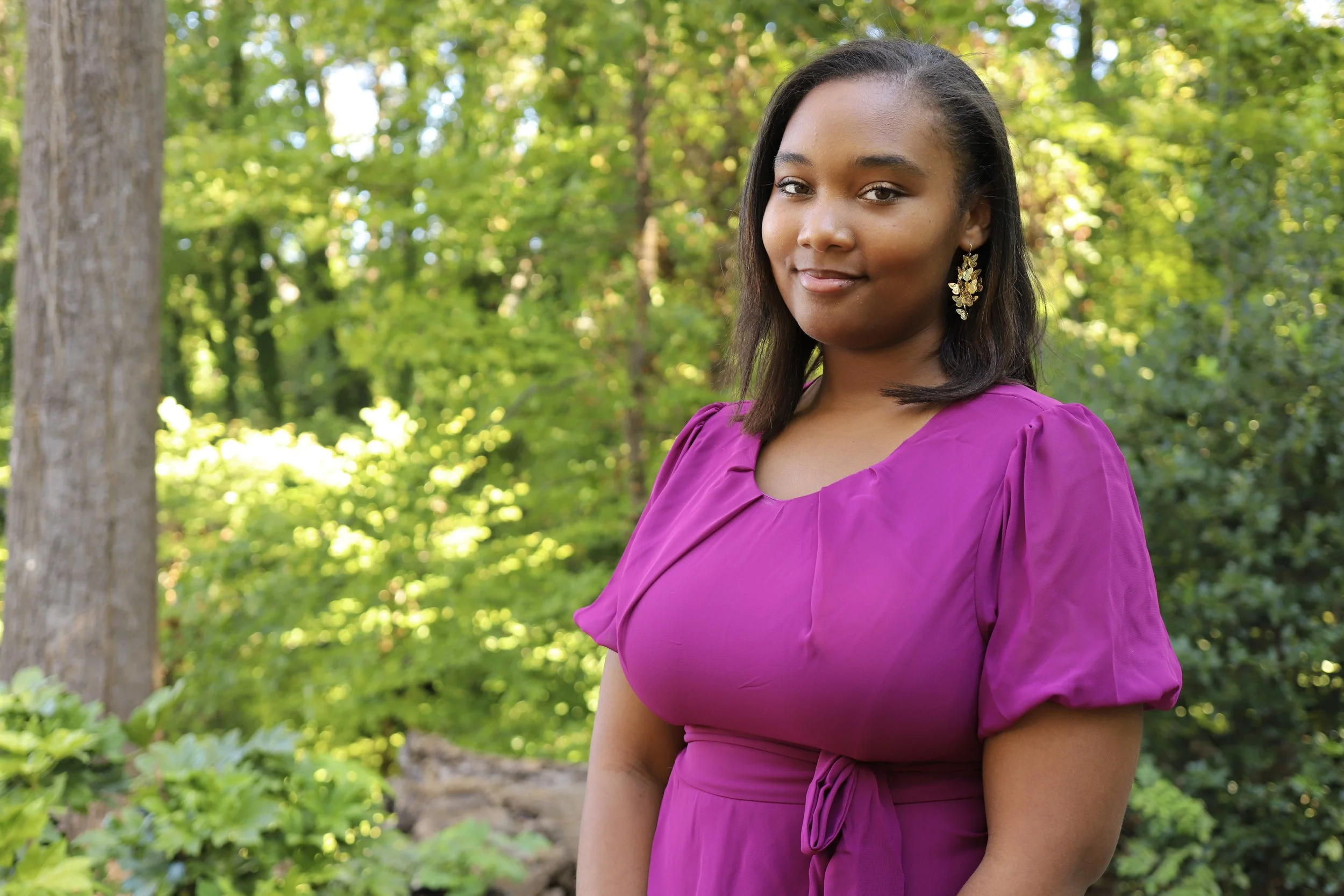 A woman in a purple dress standing outdoors with green trees and foliage in the background.