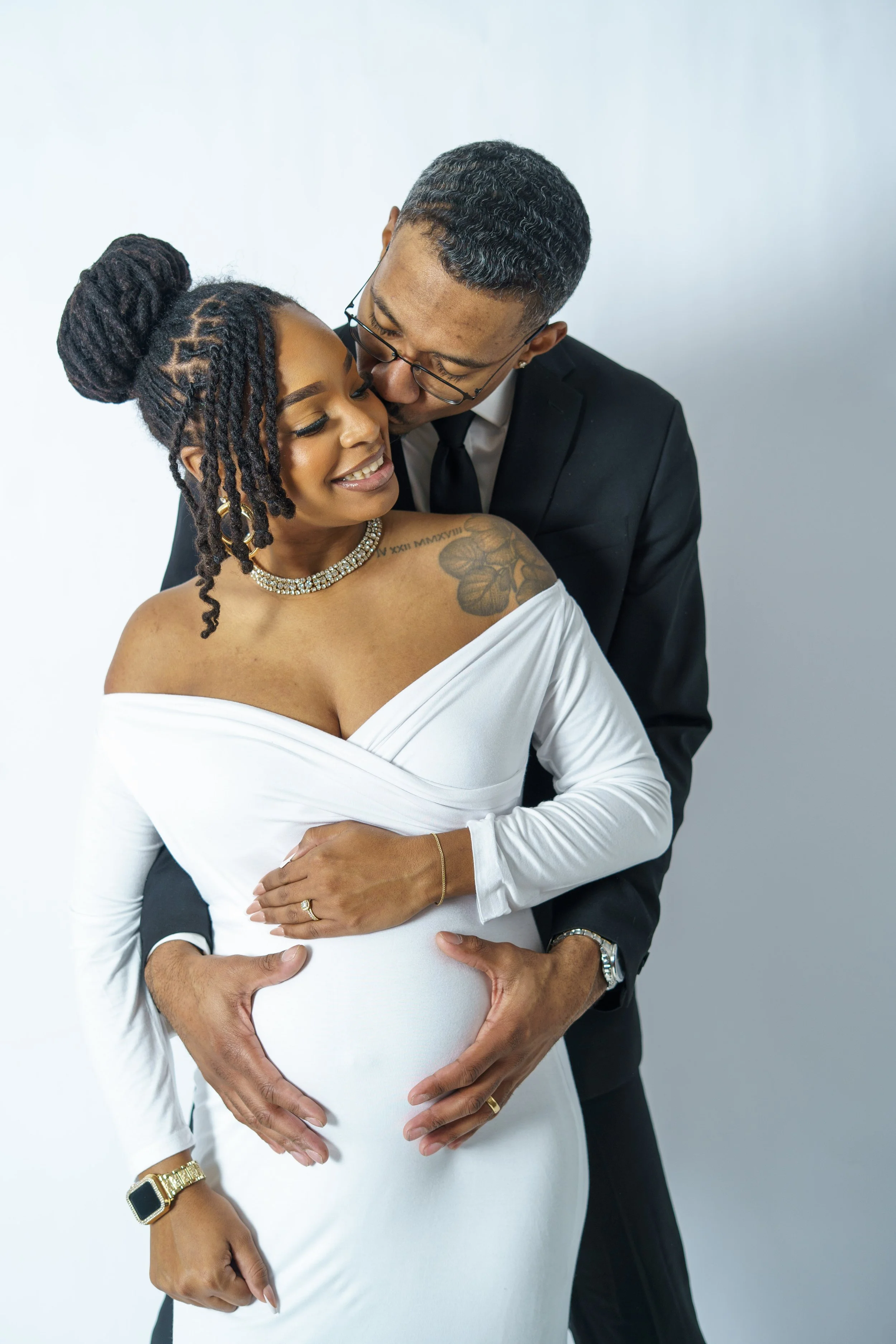 A man and woman, dressed elegantly, embrace and smile while the woman is pregnant. The woman wears a white off-shoulder dress, jewelry, and has tattoos, and the man is in a black suit with glasses, both showing affection.