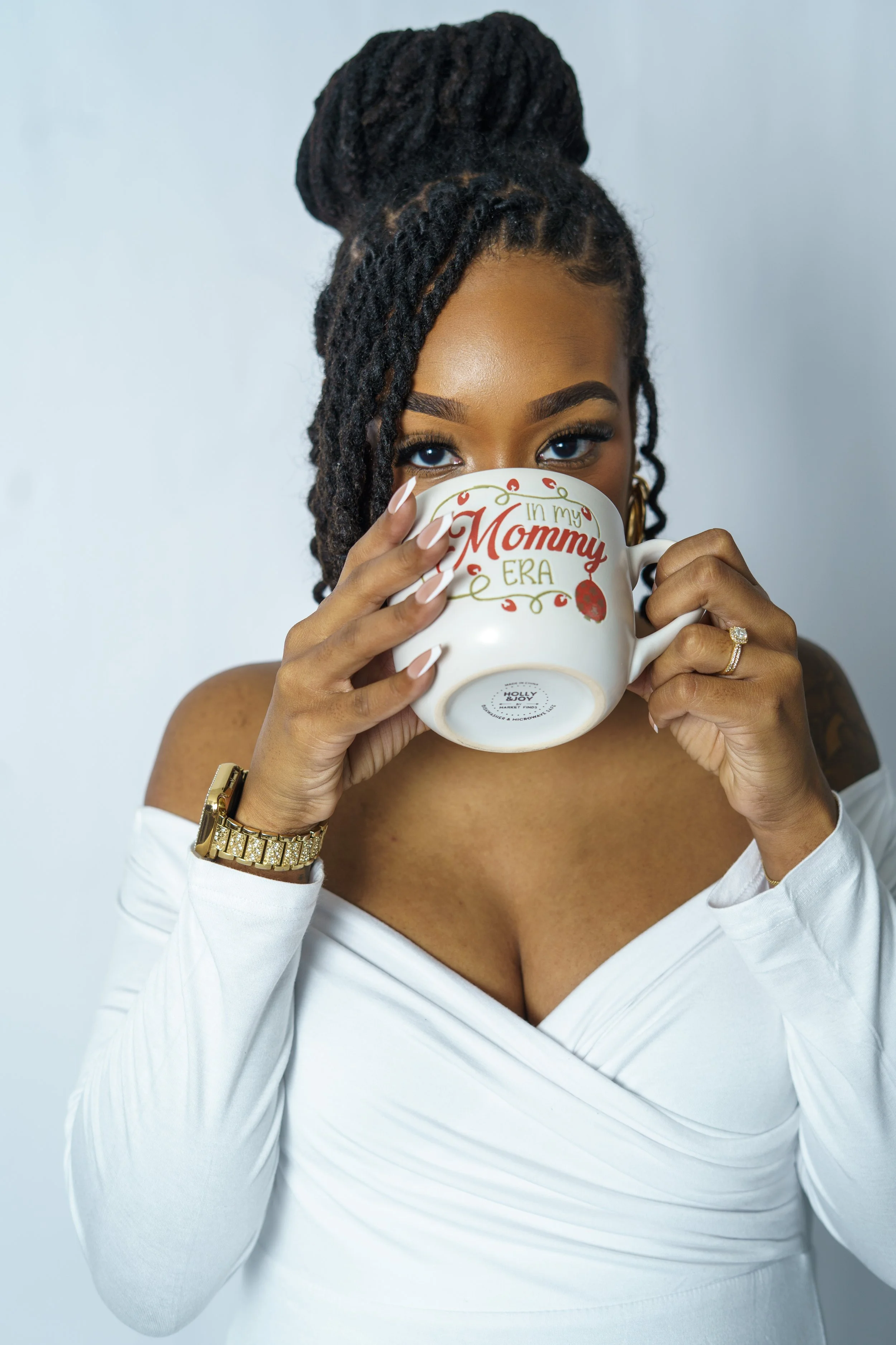 A woman with styled dreadlocks and gold jewelry holds a white mug with red and green writing that says "I'm my Mommy ERA," covering the lower half of her face, revealing her blue eyes and wearing an off-the-shoulder white top.