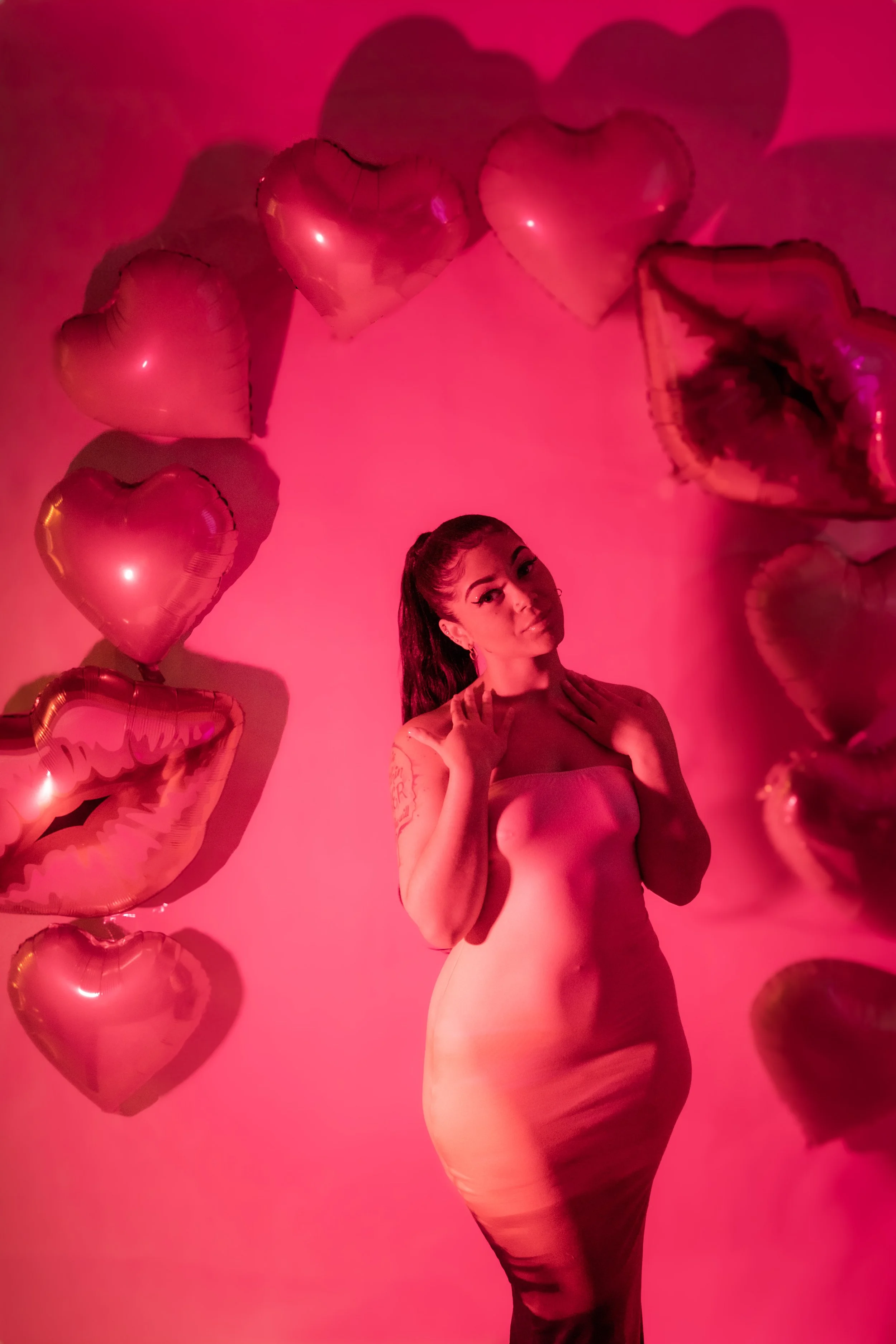 A woman in a nude-colored dress standing amidst pink heart-shaped balloons, with pink lighting creating a romantic atmosphere.