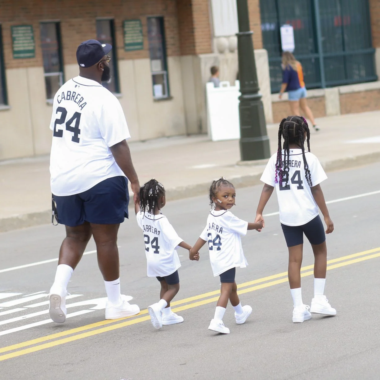 A man walking with three young girls on a city street, all wearing matching white sports jerseys with the name Cabrera and the number 24, holding hands.