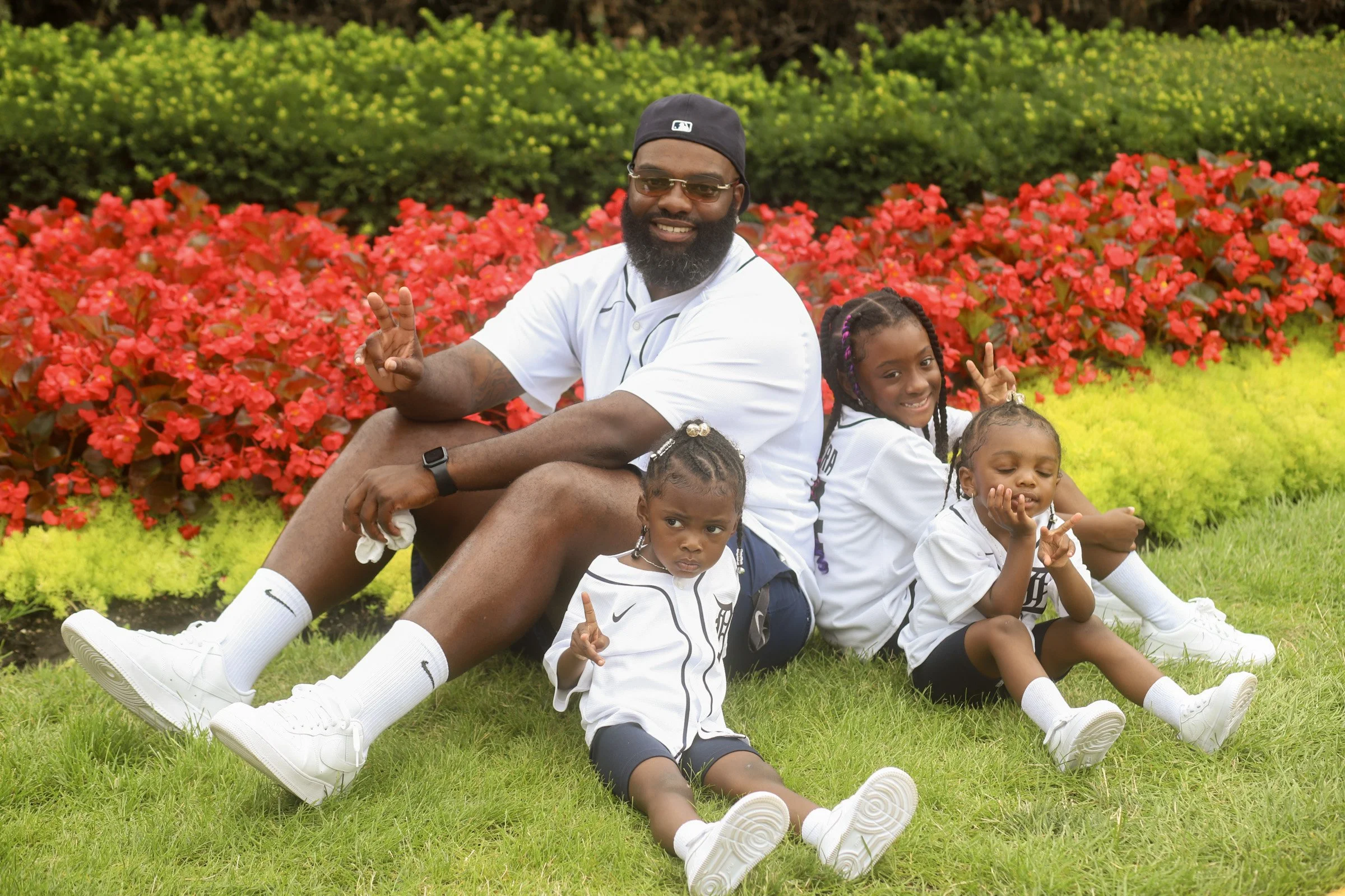 A man with three young girls sitting on grass in front of red and yellow flowers, all smiling and making peace signs.