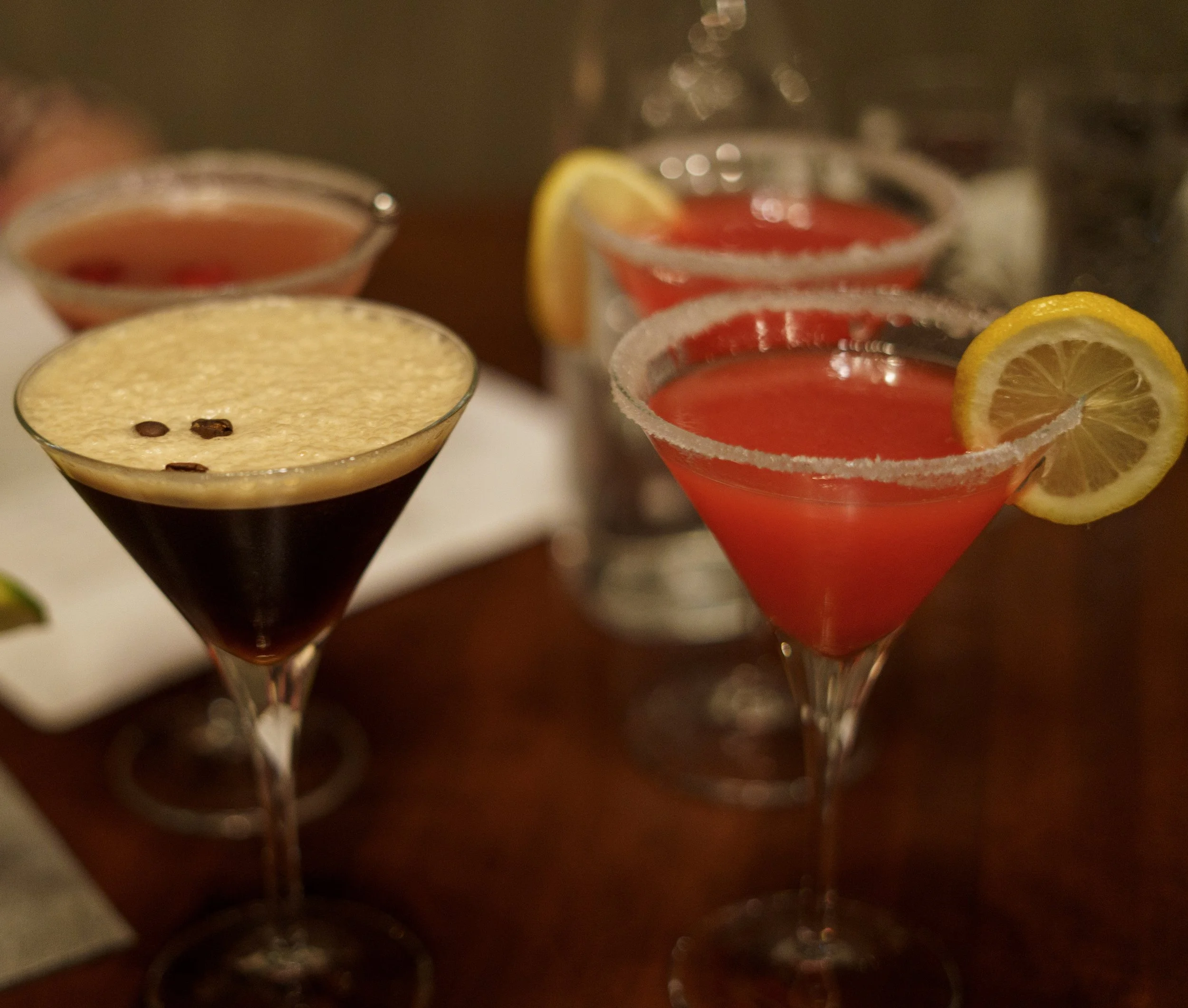 Three cocktails with lemon garnish and salted rims, one topped with coffee beans, the others with red-colored drinks, on a wooden table.