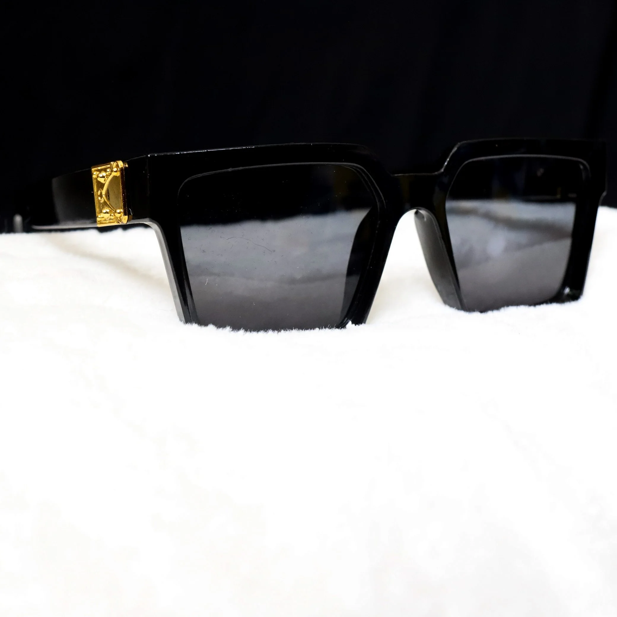 Black sunglasses with gold embellishment on the temple, resting on a white surface with a dark background.
