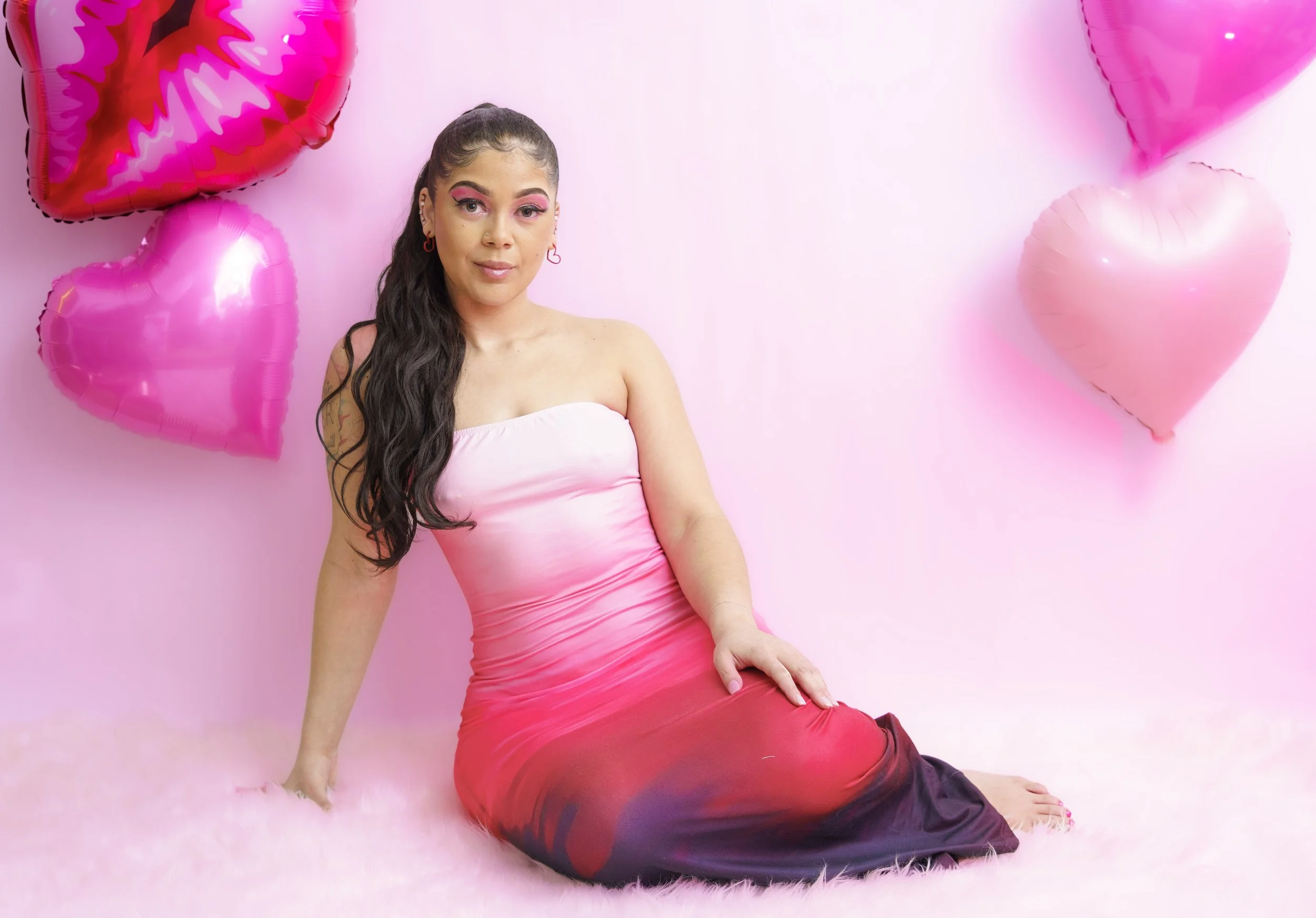 A young woman with long dark hair and pink makeup sitting on a fluffy pink rug, wearing a strapless pink and red ombre dress, surrounded by pink heart-shaped balloons against a pink background.