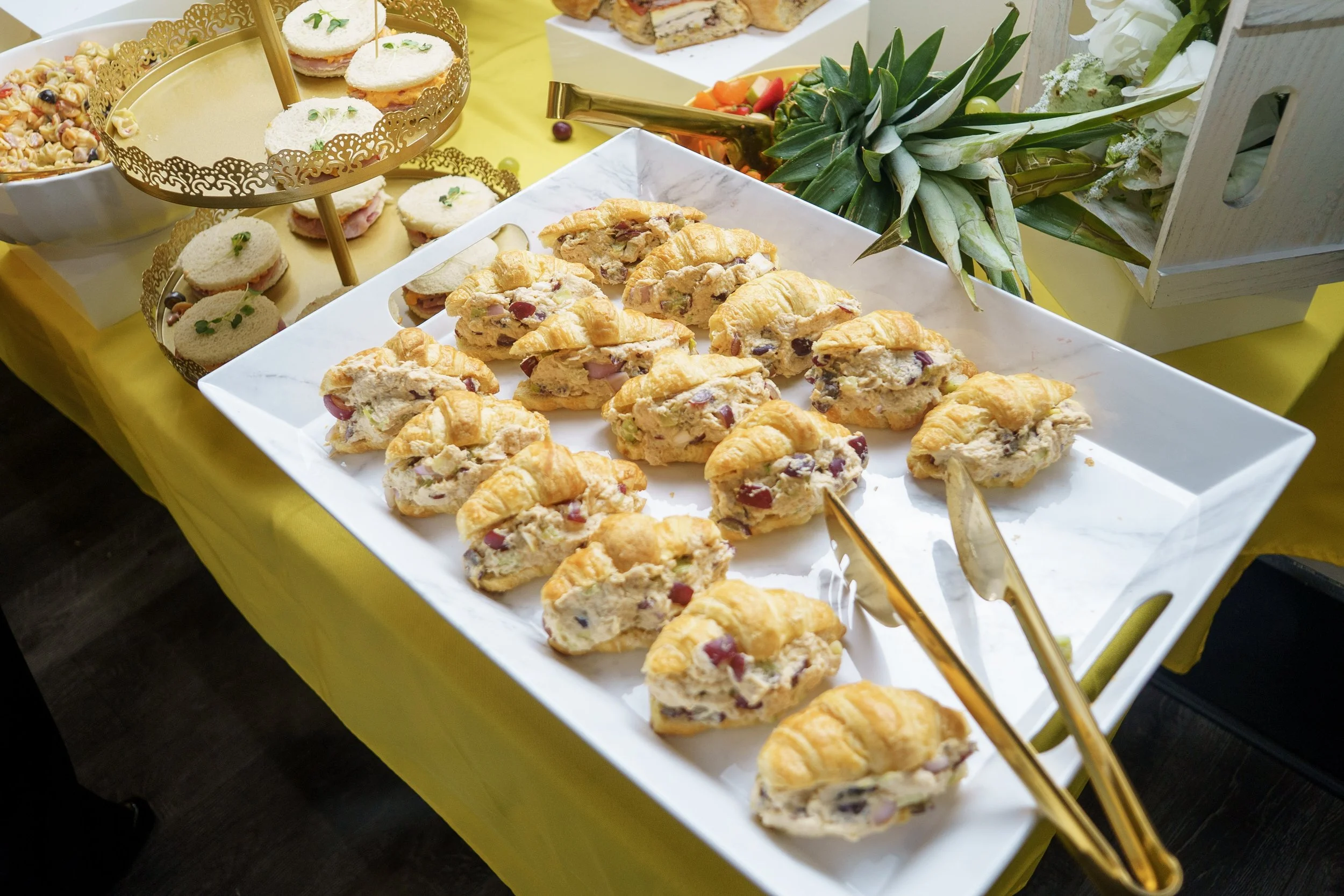 Tray of sliced croissant sandwiches with deli meats and cheese, arranged on a white platter at a buffet table decorated with a yellow tablecloth and tropical fruit.
