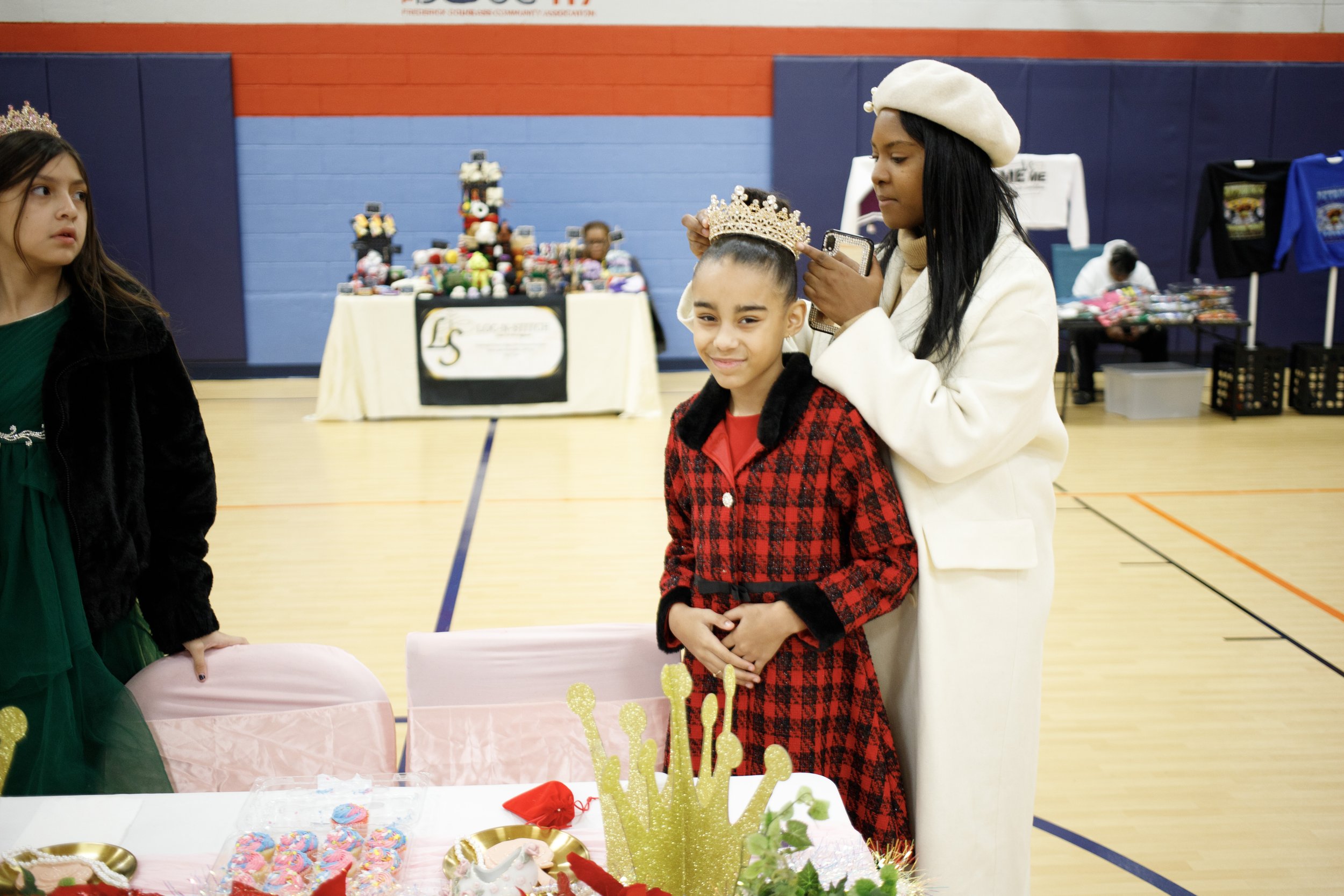 Girl wearing a crown is being celebrated at a birthday party, with another woman placing the crown on her head, as children and party decorations are visible in the background.