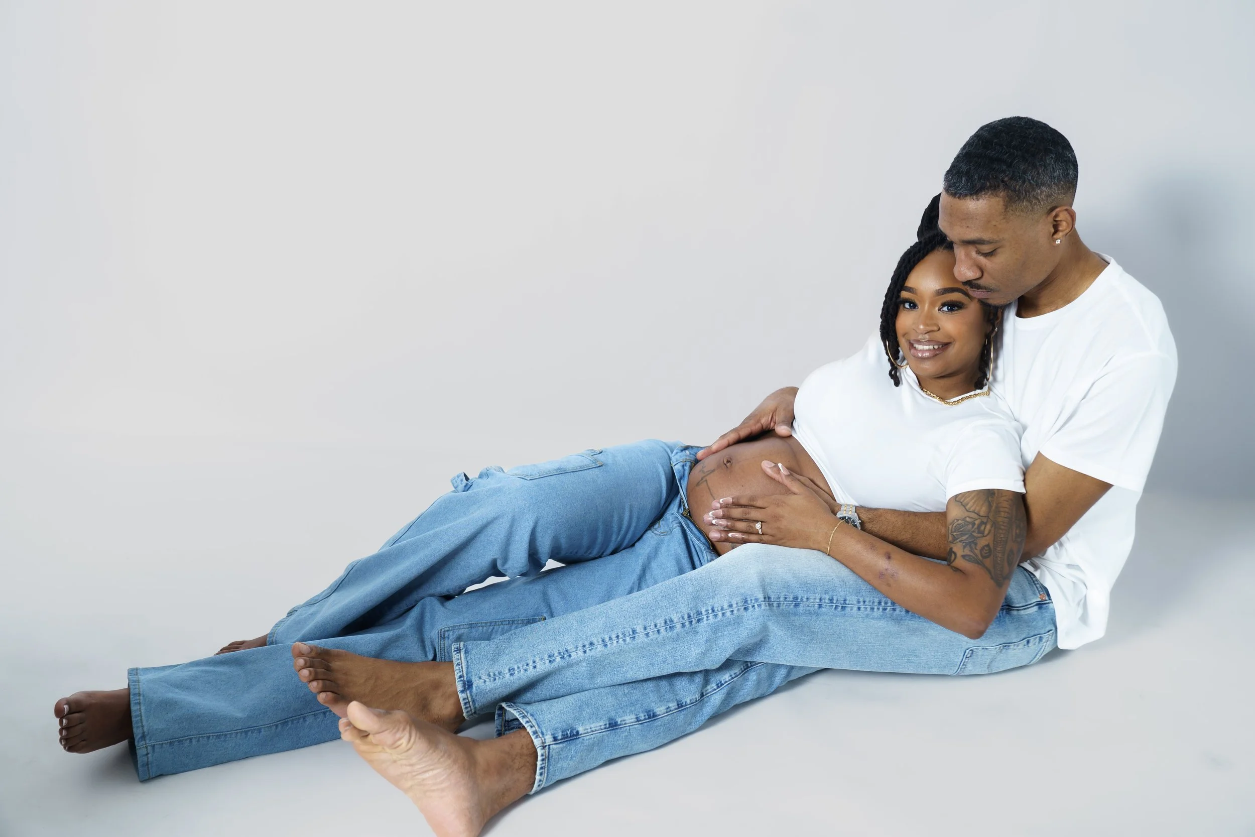 A happy pregnant woman lying on her partner's lap with the man cradling her belly, both smiling affectionately, against a plain light background.