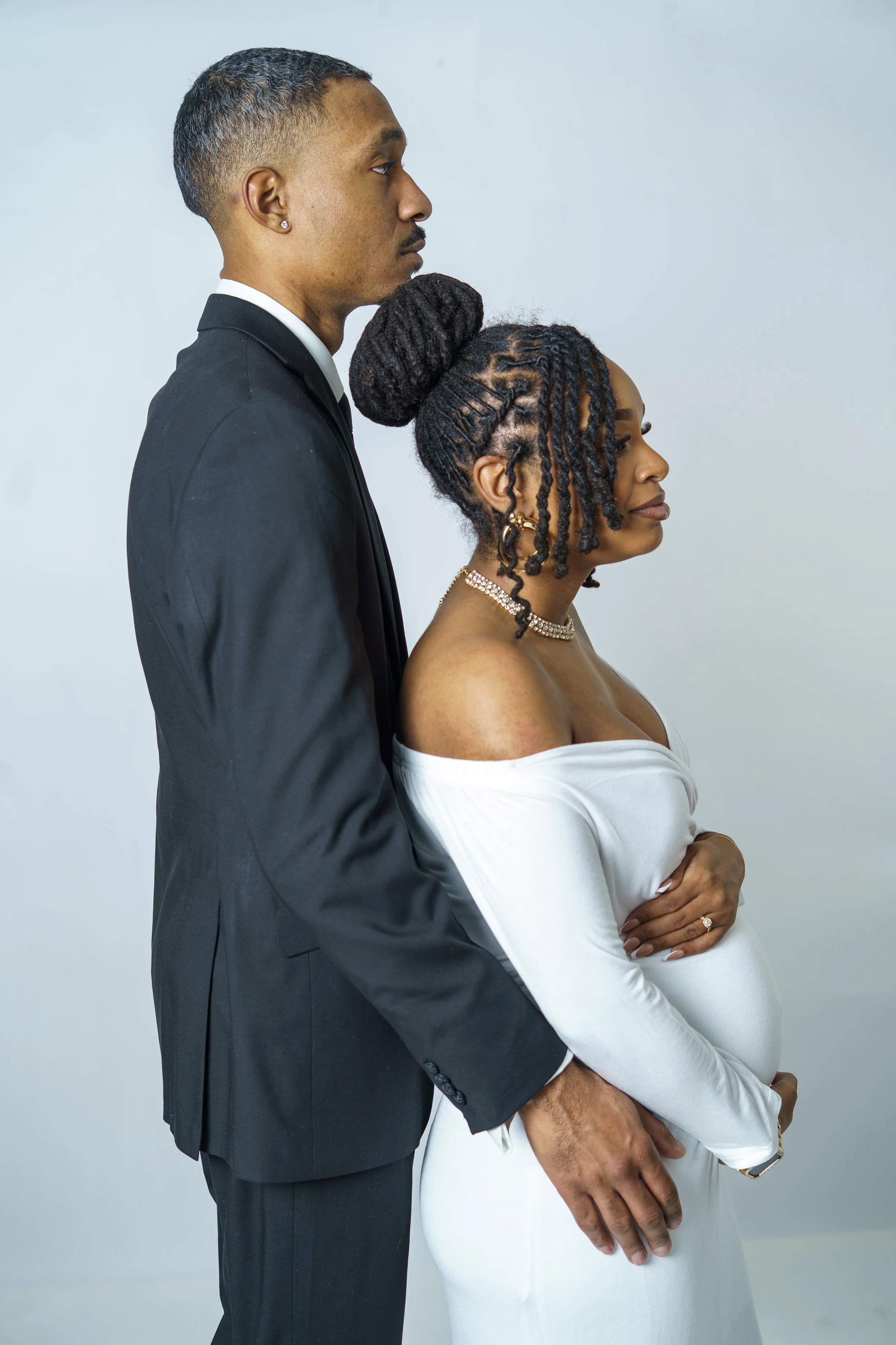 A couple, with the man dressed in a black suit and the woman in a white off-shoulder dress, standing back to back against a plain light background, with the woman gently holding her pregnant belly.