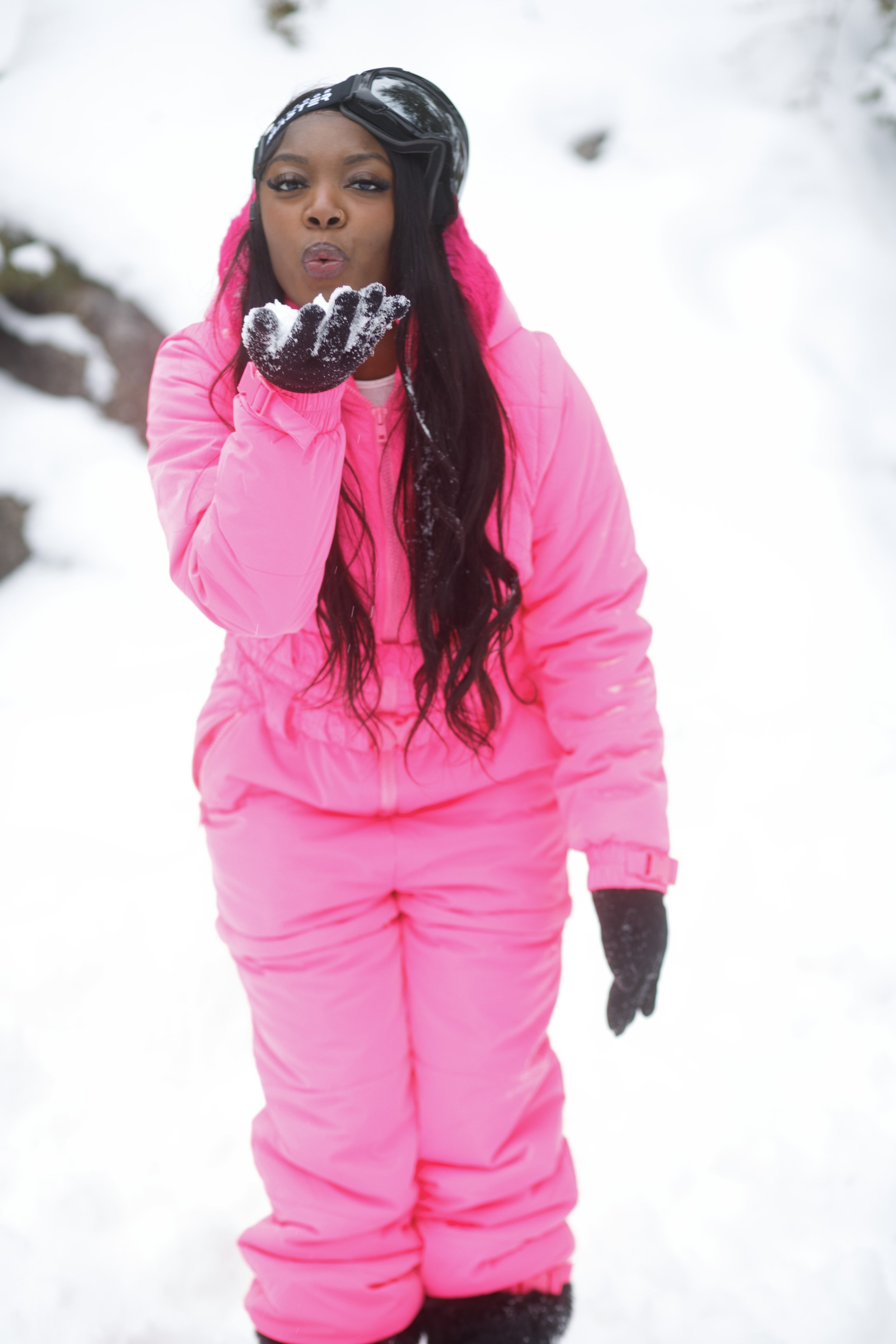 Woman in a pink snowsuit blowing snow off her gloved hand outdoors in a snowy landscape.