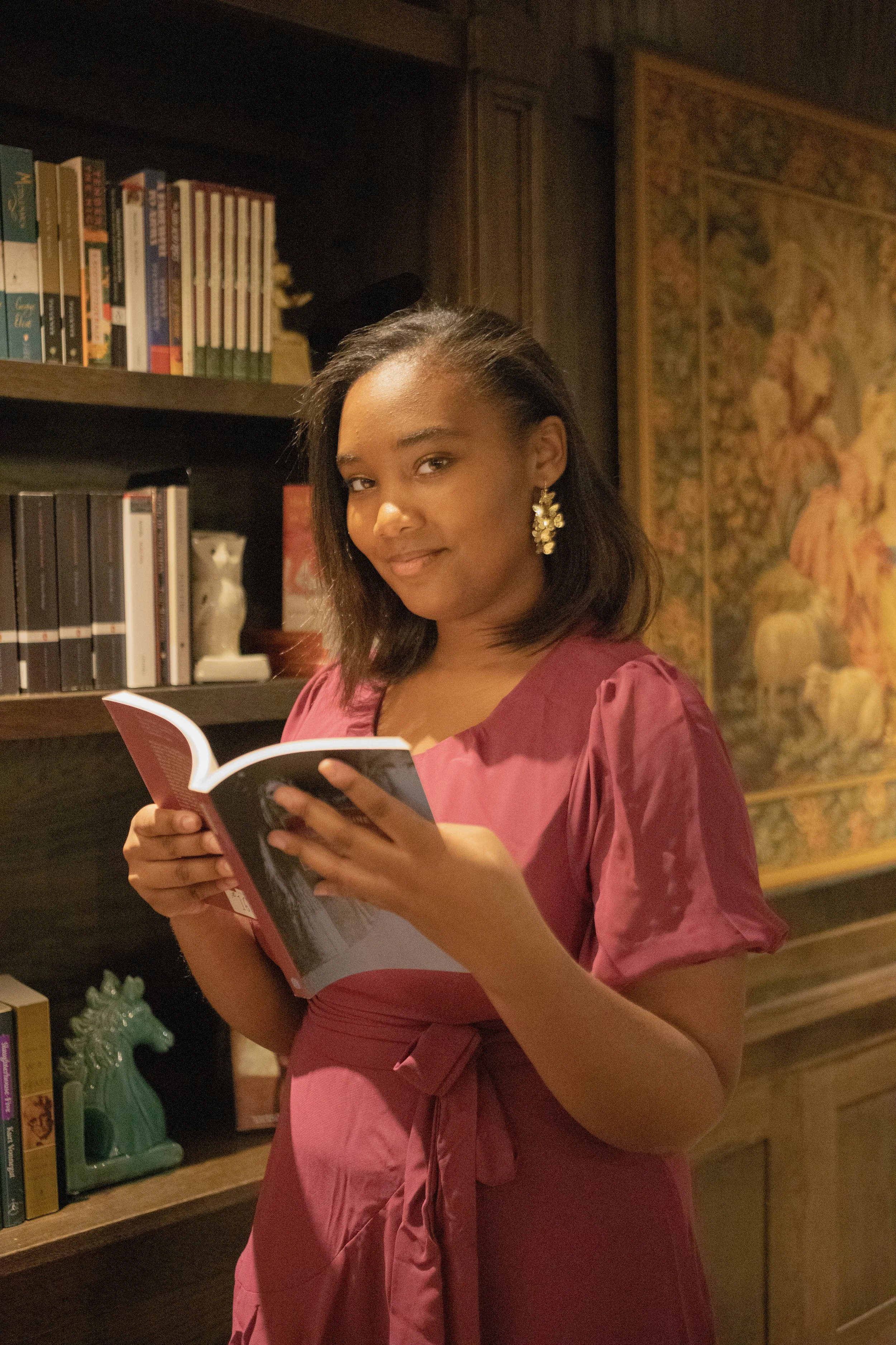 A woman in a pink dress holding a book in a library with bookshelves and a decorative wall hanging in the background.