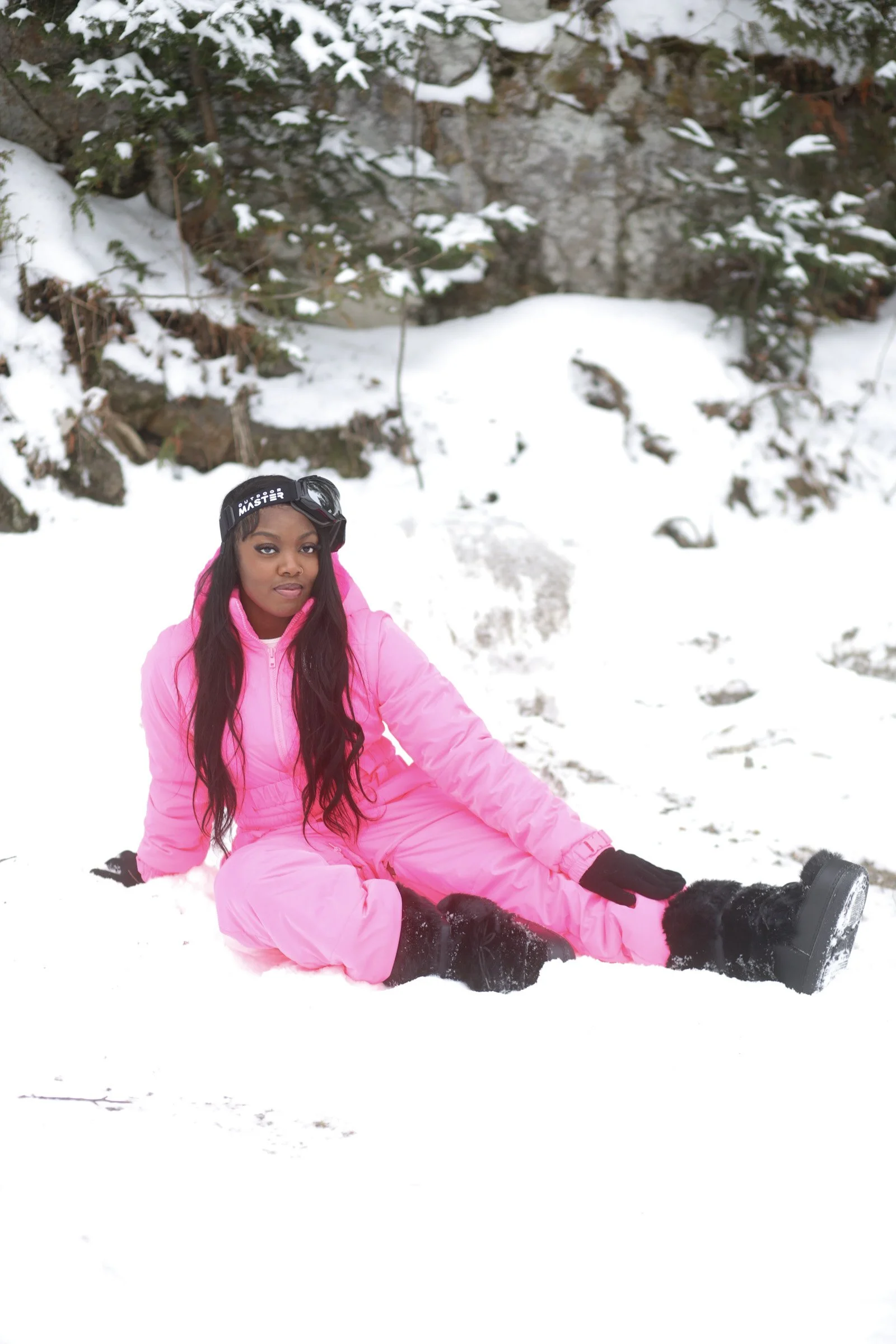 A woman in pink winter clothing sitting on snow with trees and rocks in the background.