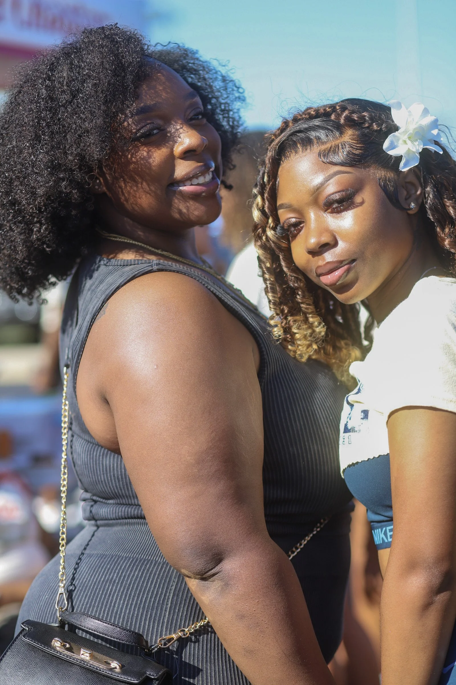 Two women standing close together outdoors during the day, one smiling and the other with a serious expression, sunlight casting shadows on their faces.