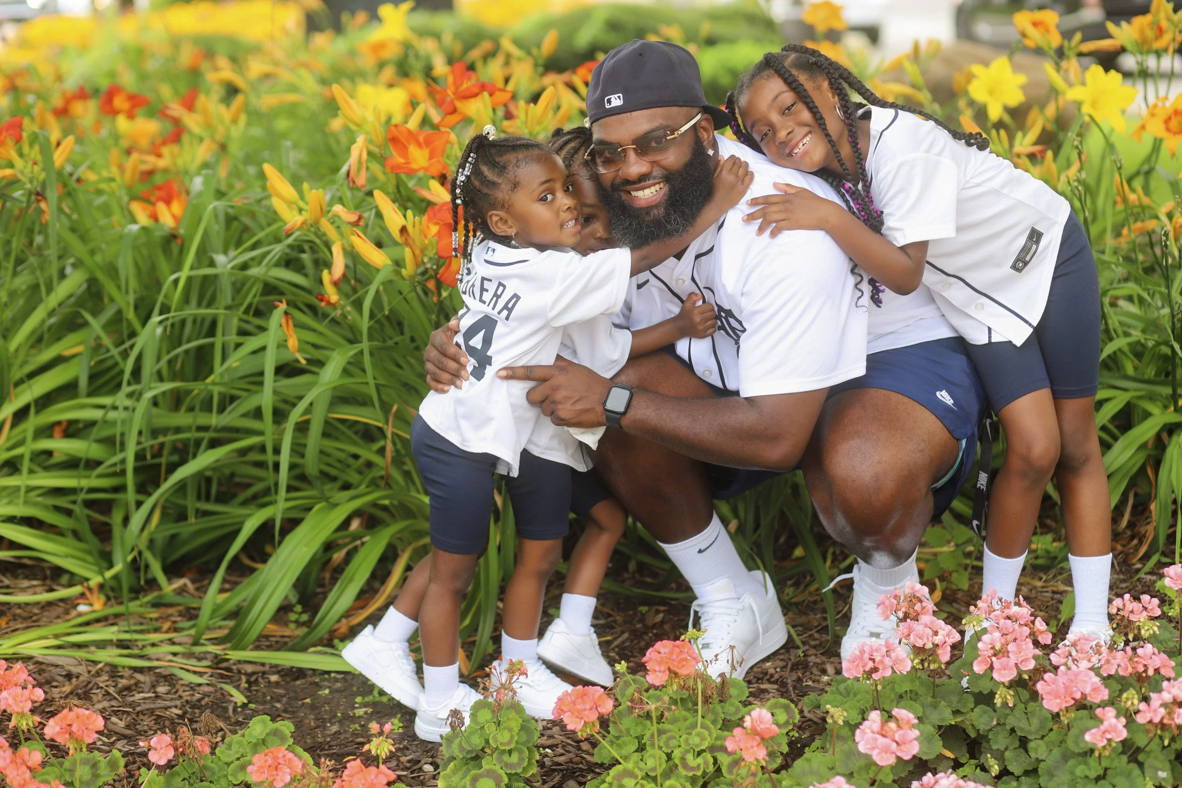 A joyful family of five, including a man, woman, and three young girls, wearing matching white and navy sportswear, hugging and smiling in a colorful garden filled with orange, yellow, and pink flowers.