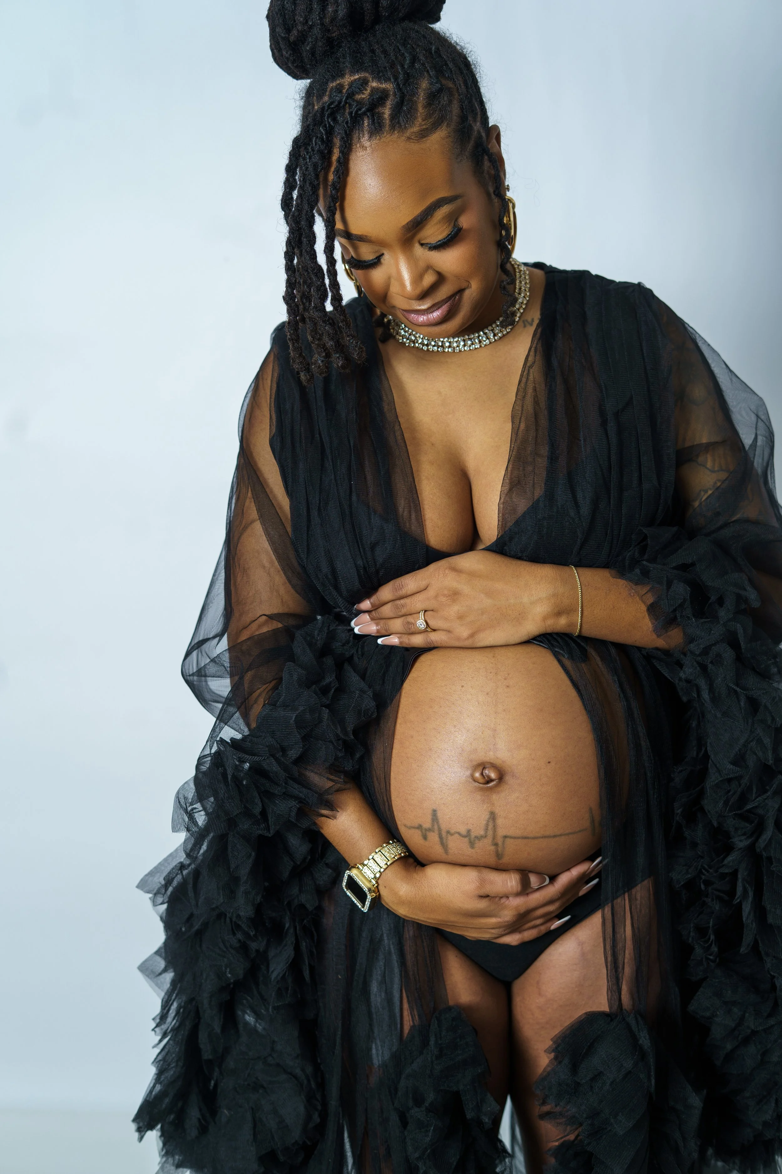 A pregnant woman in a sheer black gown with ruffle details, smiling gently, holding her belly which has a heartbeat tattoo, against a plain background.