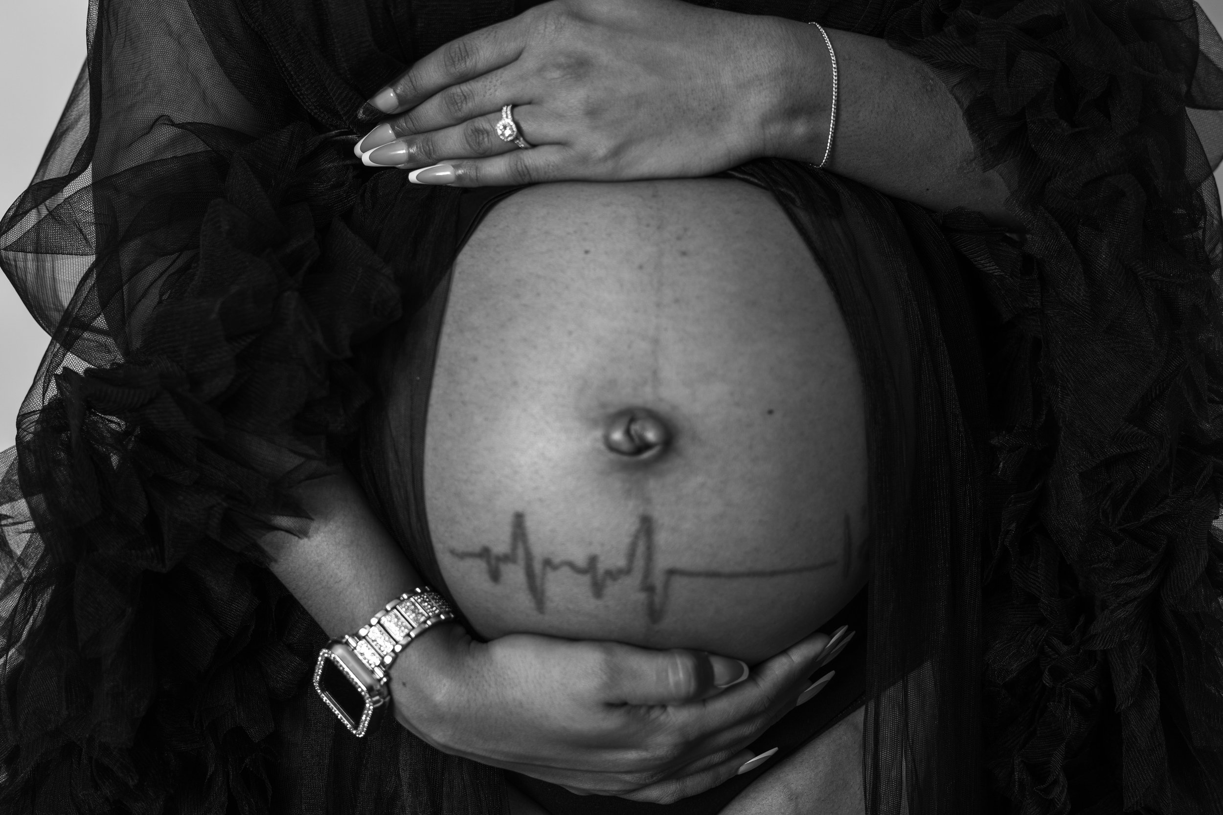 Close-up of a woman with a tattoo of an EKG line on her stomach, holding her exposed belly while wearing a ring, bracelet, and a black ruffled garment.