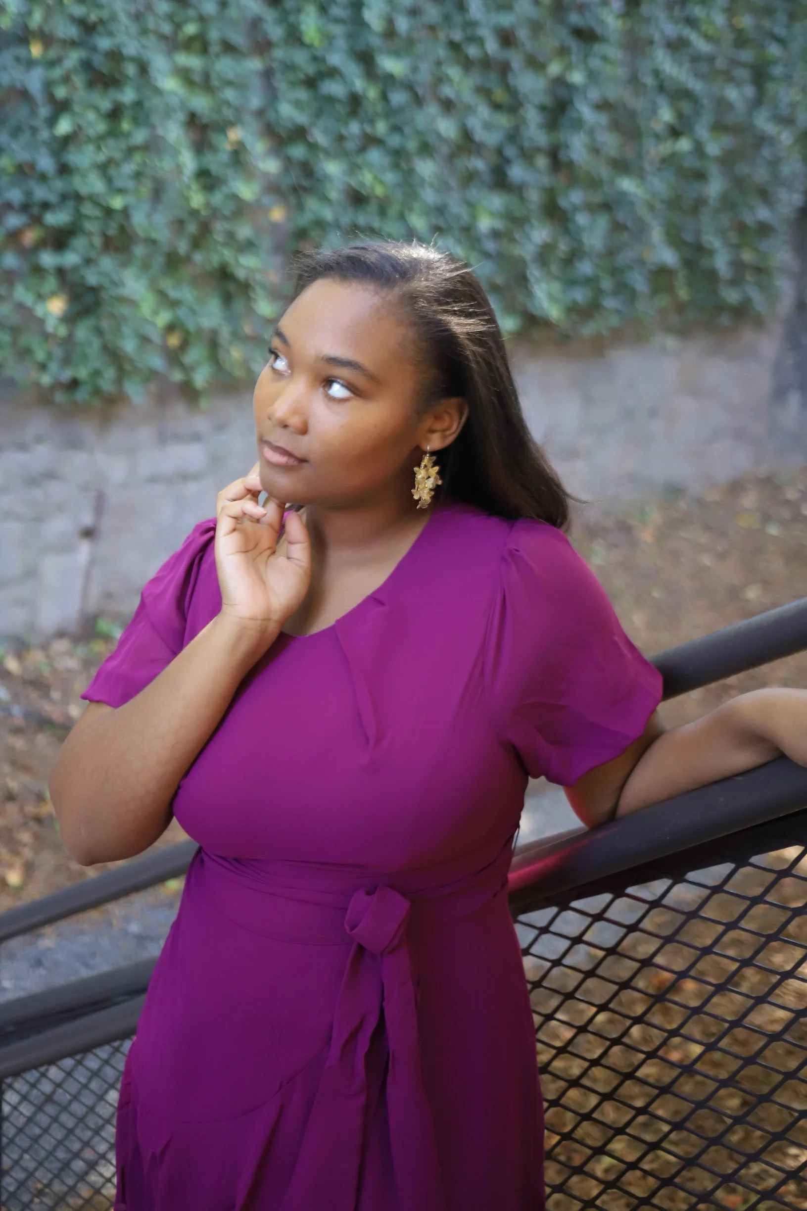 A woman with medium skin tone wearing a purple dress and gold earrings standing outdoors by a black railing, with green foliage and a stone wall in the background.