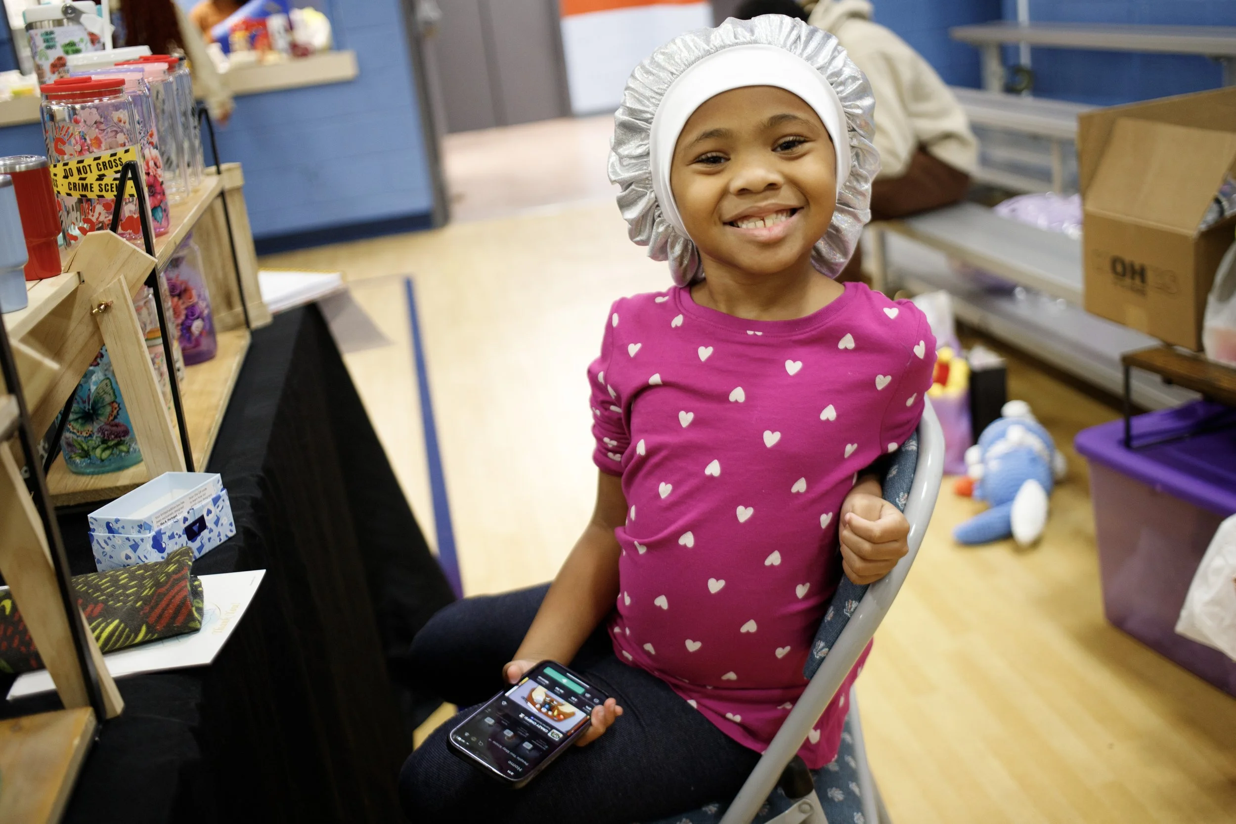 A young girl smiling while sitting on a chair in a craft or toy store, wearing a pink shirt with white hearts, a white hair cap, and holding a smartphone with a game or app open.