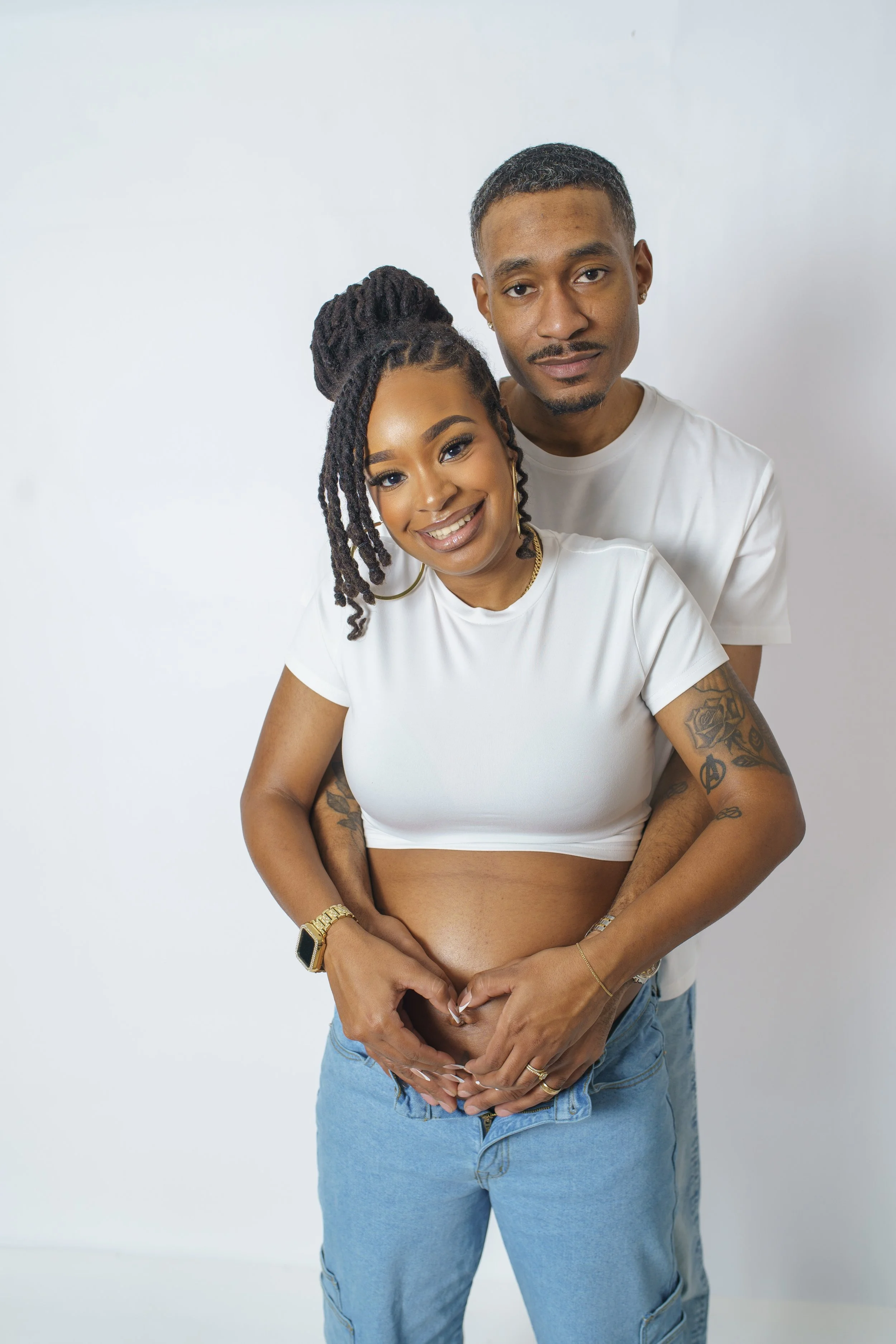 A happy pregnant woman with curly hair and a man with short hair and a mustache, both smiling and standing close together against a plain white background. The woman is wearing a white crop top and jeans, and the man is wearing a white t-shirt.