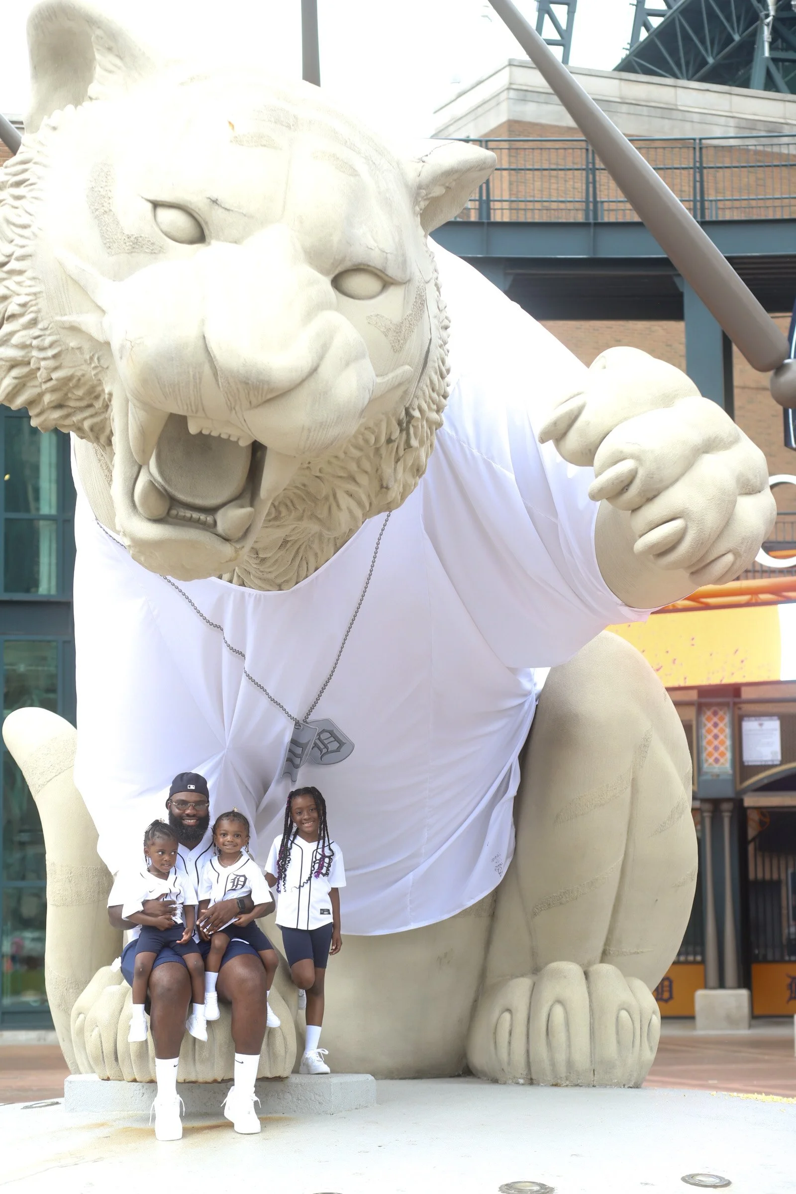 A man and three children sitting on a lion statue dressed in Detroit Tigers baseball jerseys, with a person in a lion mascot costume standing behind them.