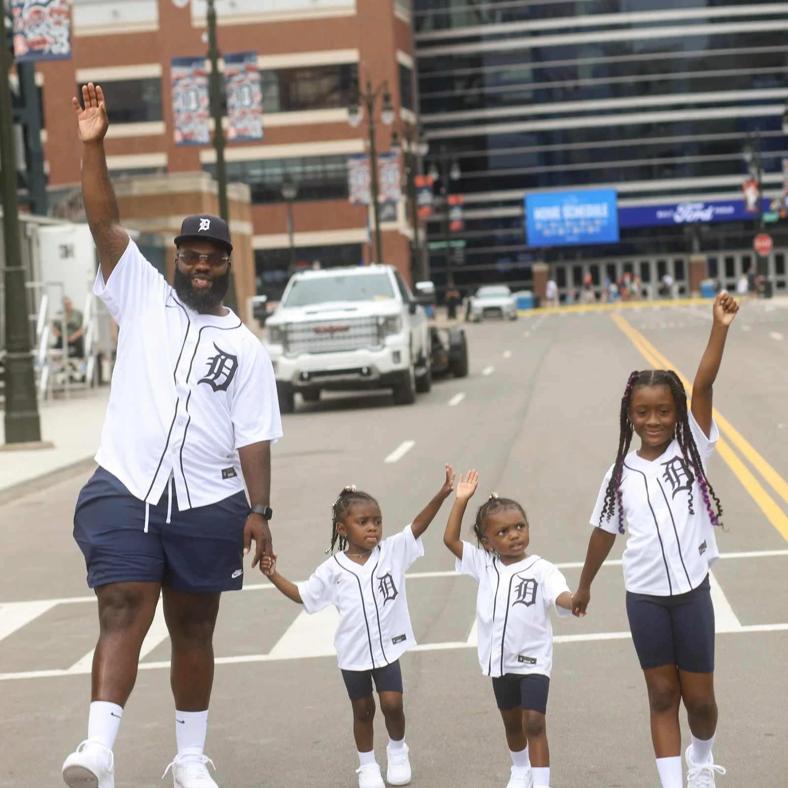 A man and three young girls, all dressed in Detroit Tigers baseball jerseys, walking hand in hand on a city street. They are smiling and raising their arms in victory or celebration.