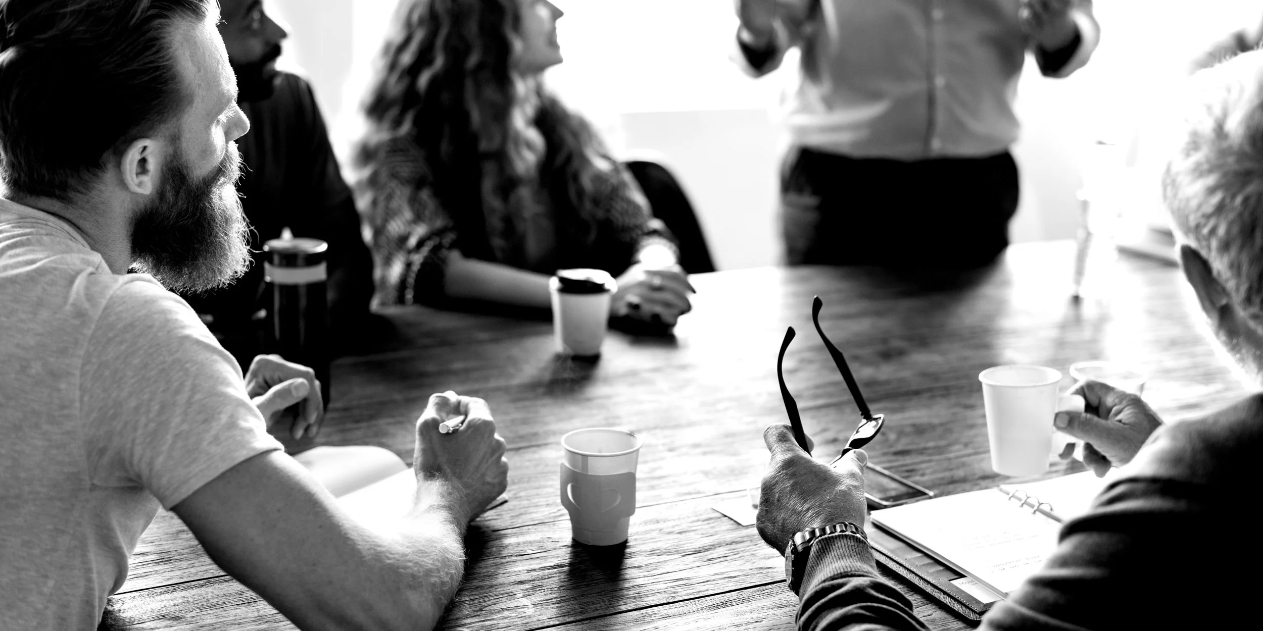 Black and white photo of a group of people sitting around a wooden table having a meeting, with cups and a notebook visible. One person is holding a pair of glasses.