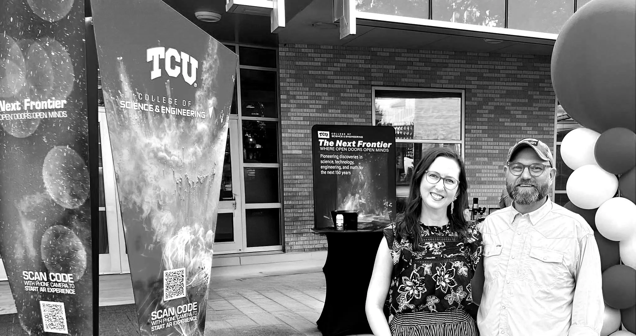 Two people, a woman and a man, standing side by side outside a building in front of a promotional display for TCU College of Science & Engineering. The woman has dark, wavy hair, glasses, and is wearing a floral top and striped skirt. The man has a beard, glasses, a cap, and is wearing a light-colored button-up shirt. There are balloons on the right side and a large sign with TCU branding and campus information in the background.