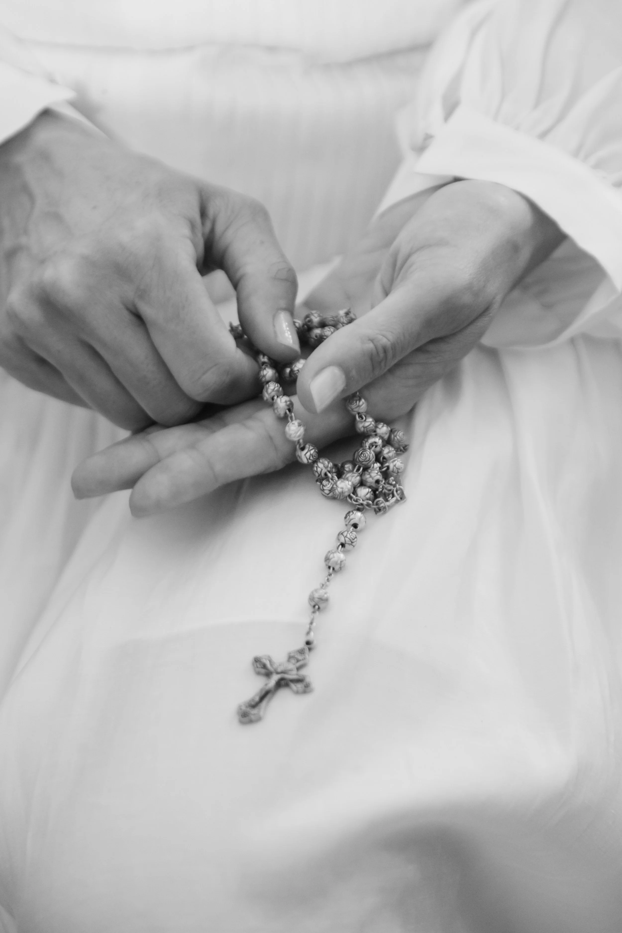 Mãos de uma pessoa segurando uma oração com um rosário de contas com uma cruz na ponta, em preto e branco.