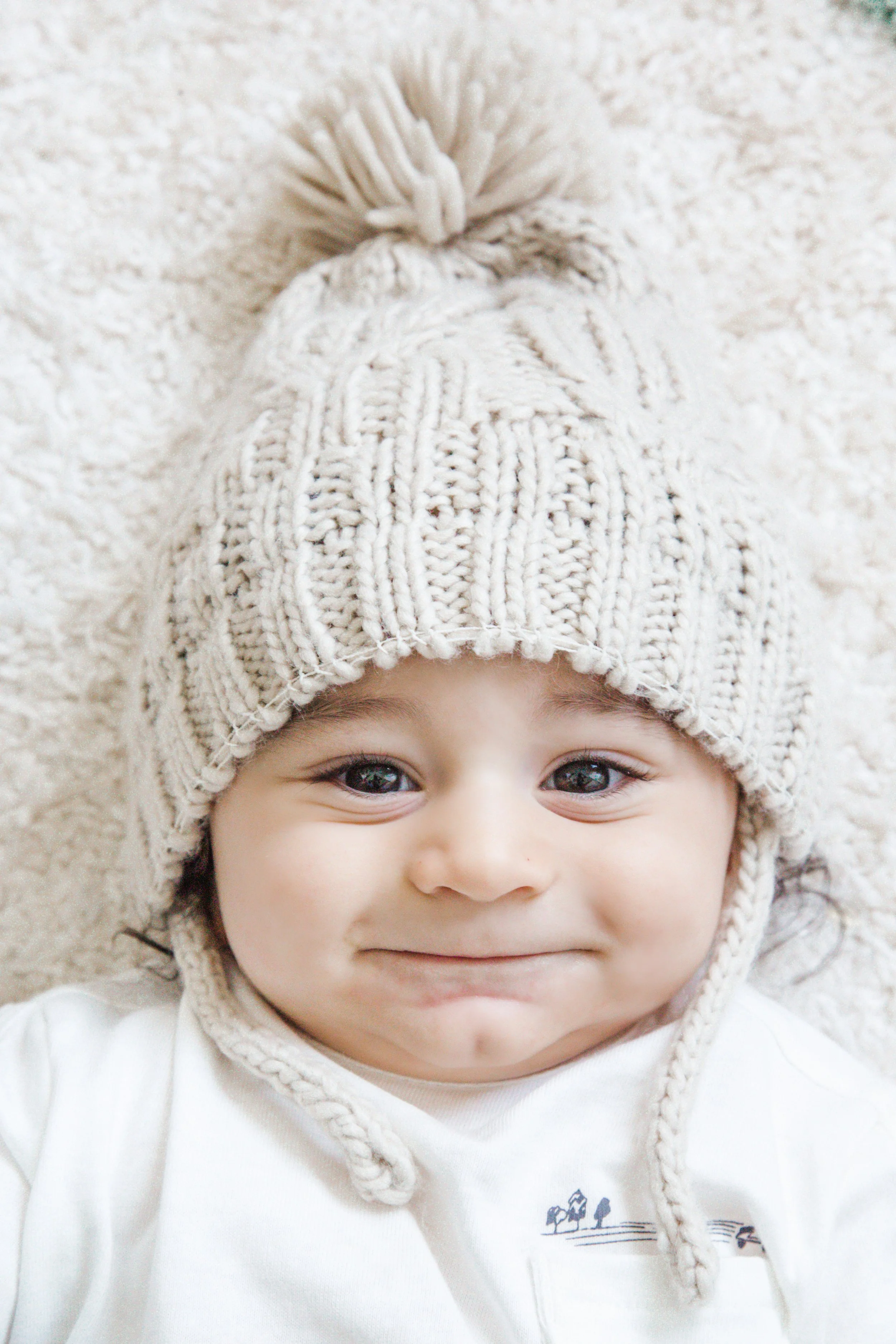 Criança de expressão tranquila, usando gorro de lã branco com pompom, deitado sobre uma superfície clara.