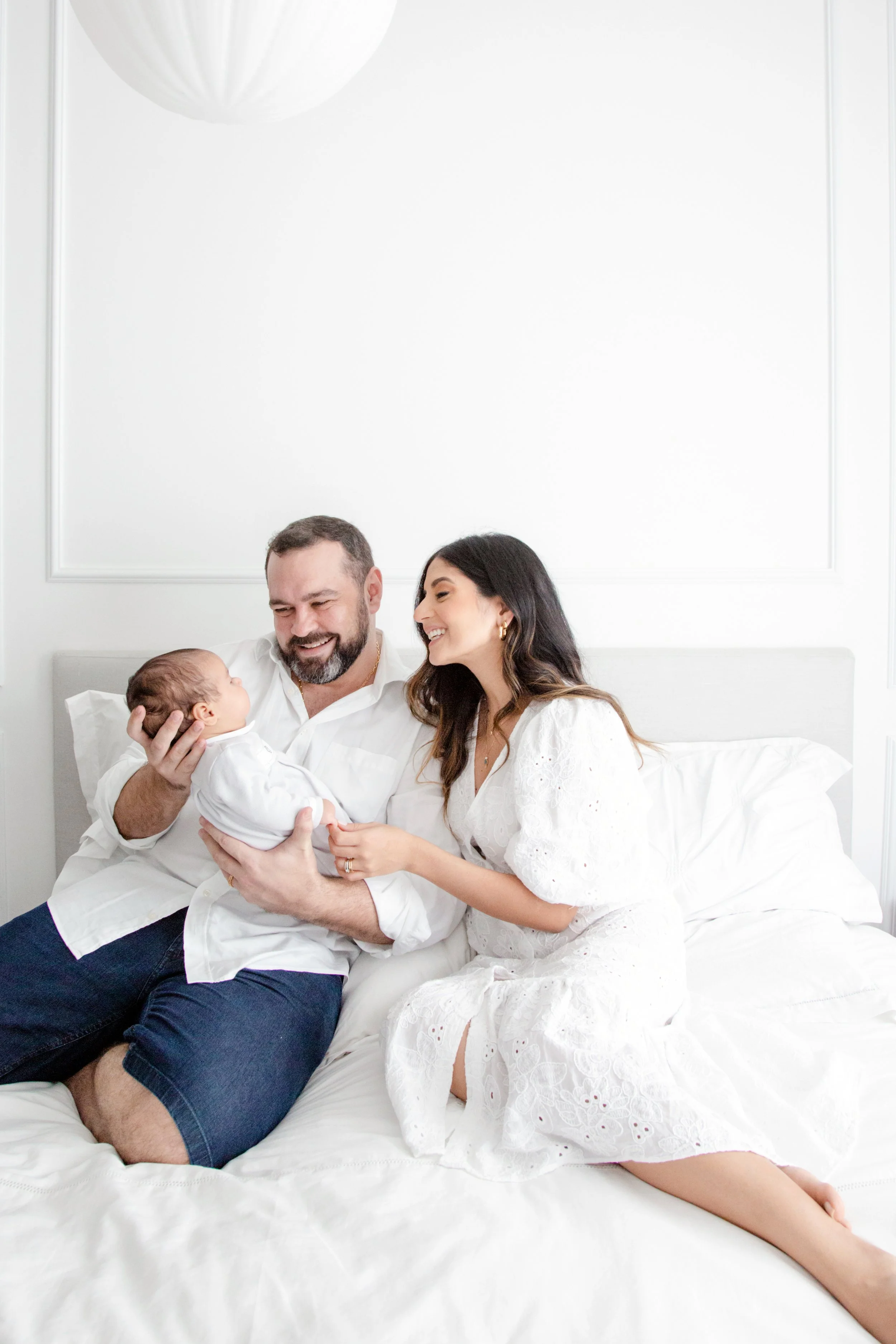 Família feliz com recém-nascido, sentada na cama, com fundo branco e decoração minimalista.