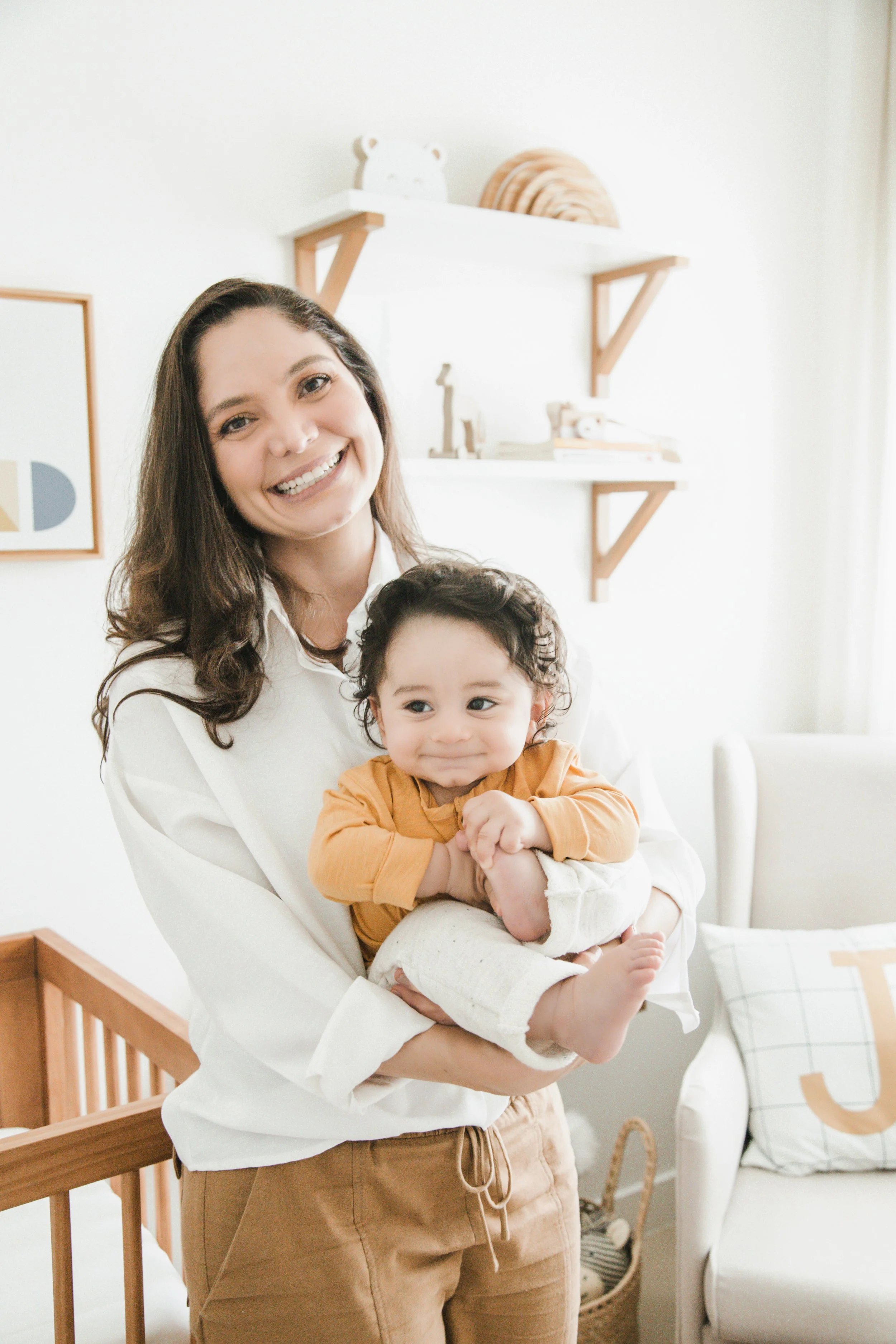 Mulher mulher sorridente segurando uma criança sorridente, dentro de uma sala decorada com objetos de madeira e uma almofada com a letra J.