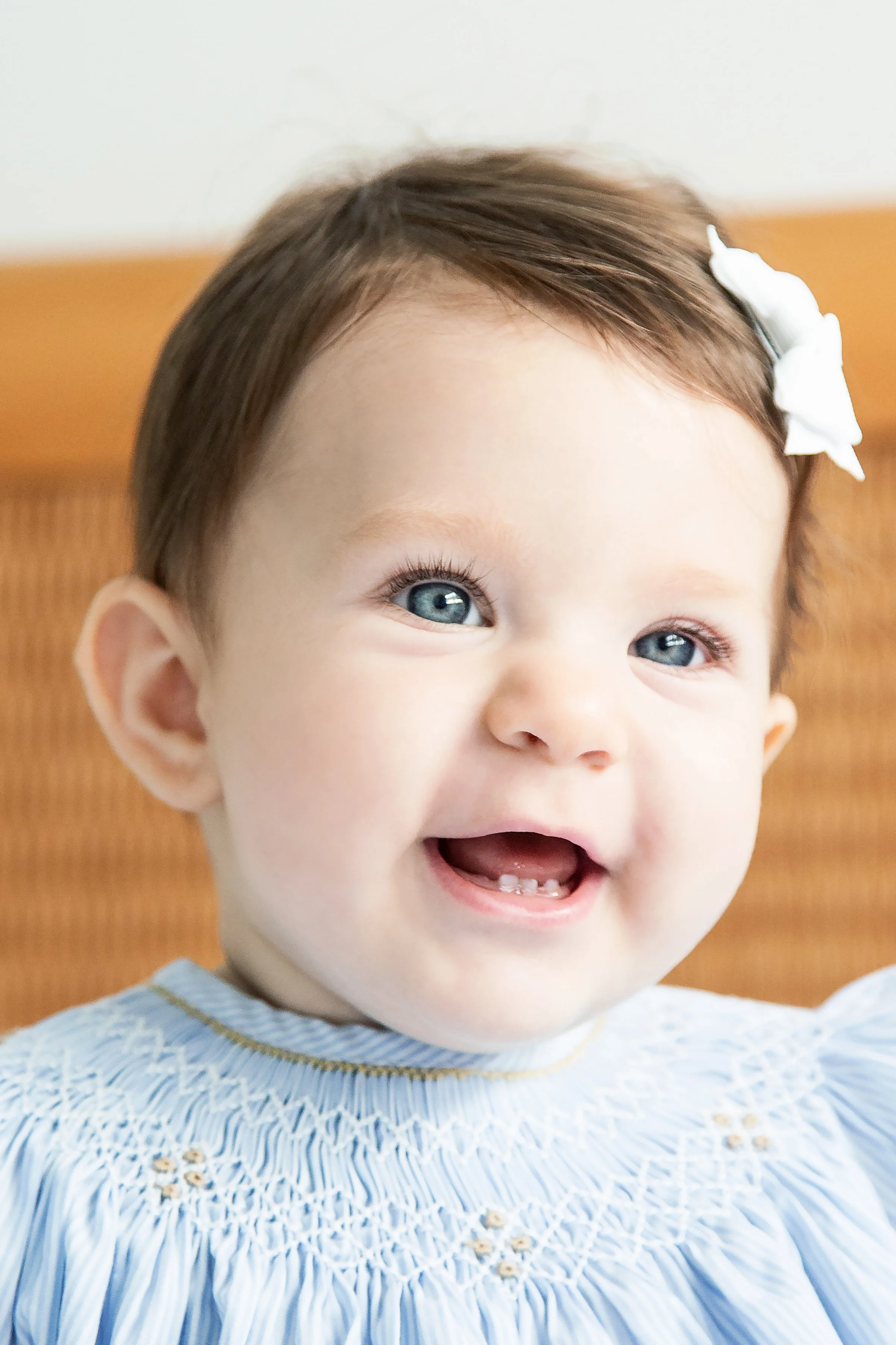 Bebê sorrindo com cabelo castanho, olhos azuis e uma cobertura branca na cabeça, vestindo blusa azul clara com detalhes crochetados.
