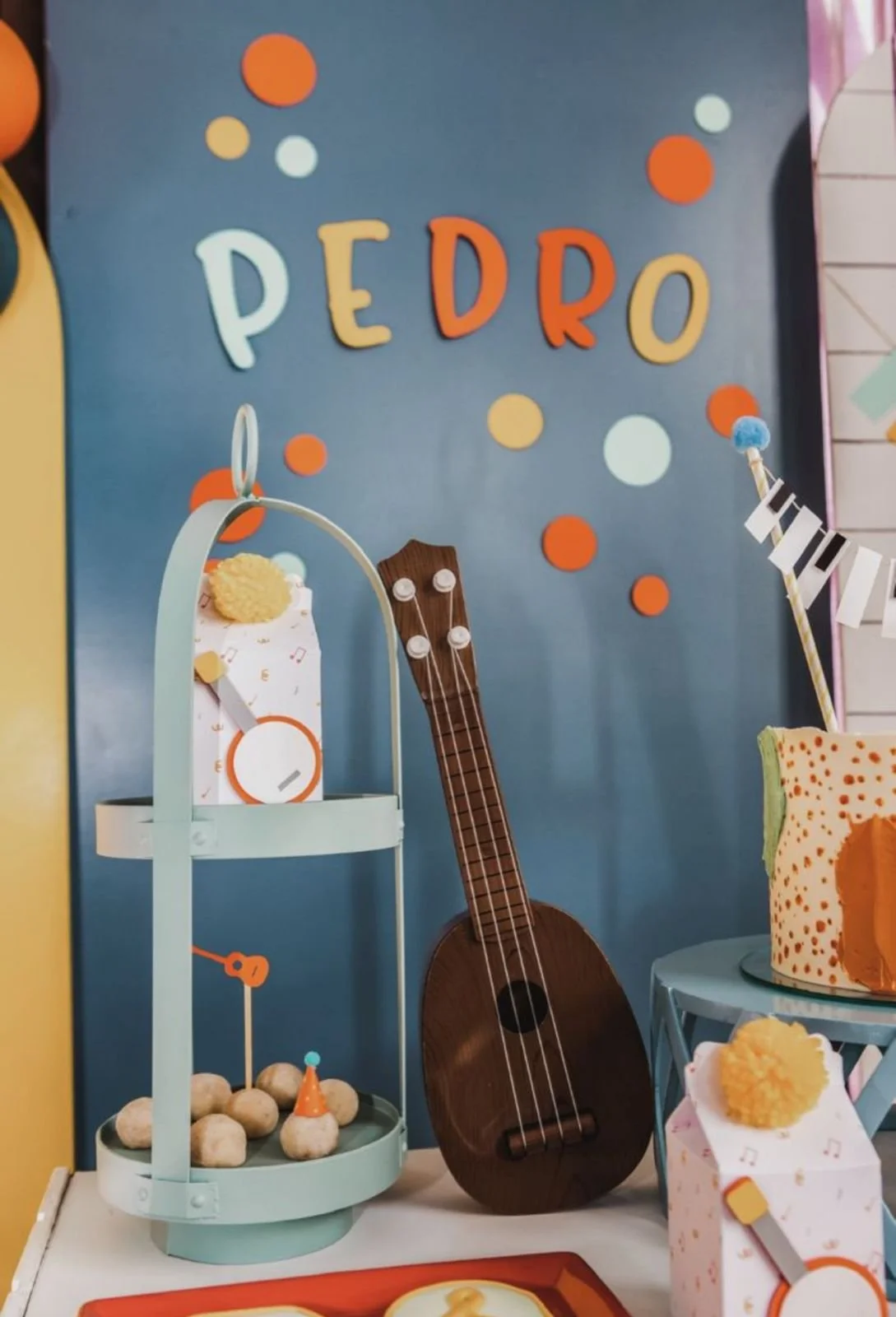 Decoração de aniversário com tema musical, com o nome Pedro em letras coloridas na parede, uma guitarra de madeira, um bolo decorado e caixas de doces.