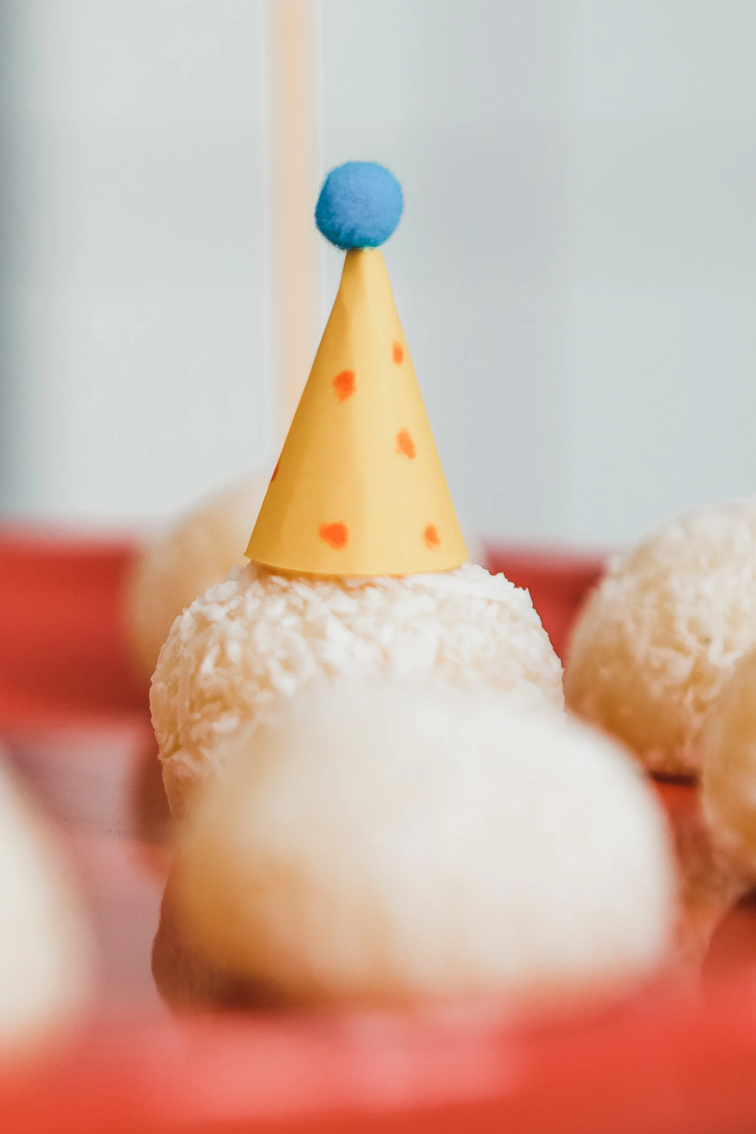 Doces decorados para aniversário com bolinha azul no topo, um deles com chapéu de festa amarelo com manchas vermelhas, e ao fundo outros doces brancos em um prato
