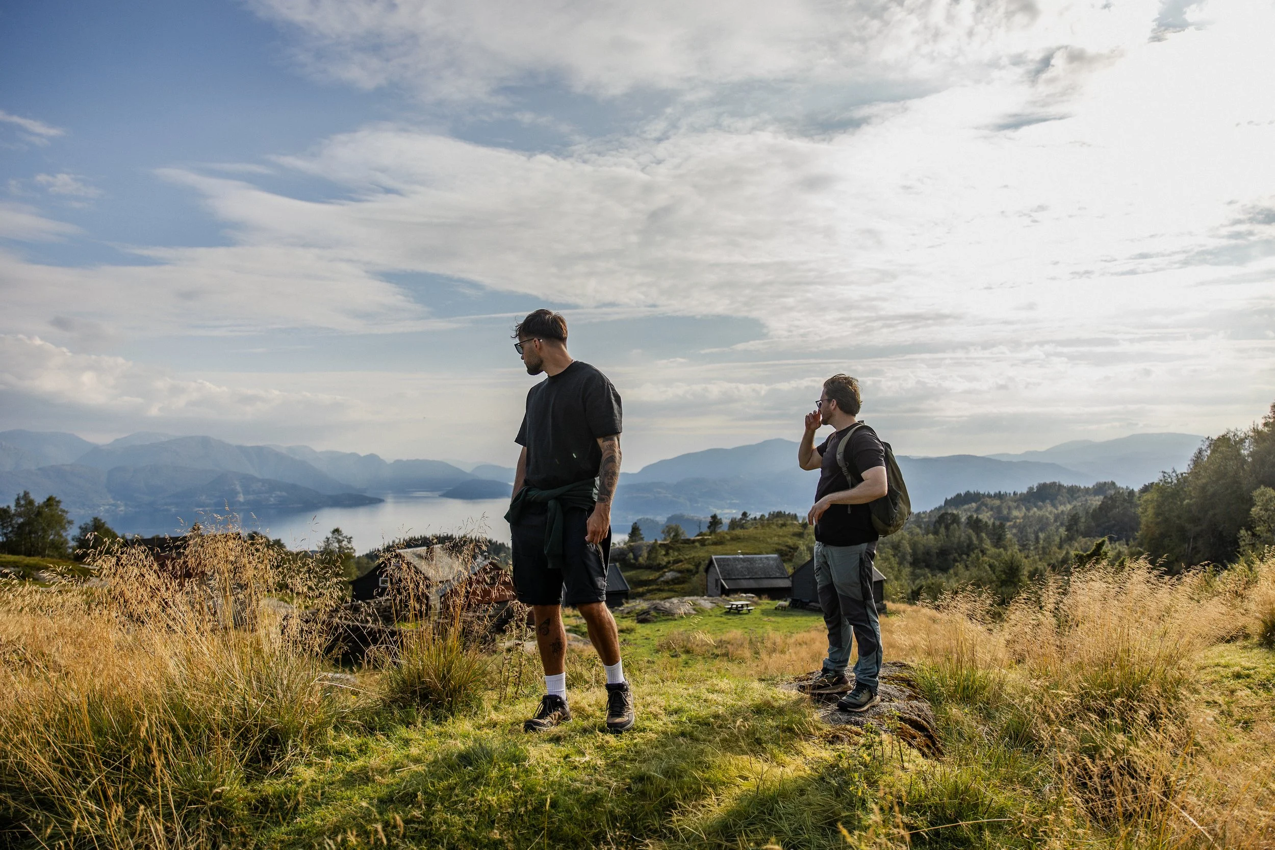 Two young men are standing outdoors in a scenic mountain landscape, one looking down and the other on the phone, with mountains, trees, and a cloudy sky in the background.