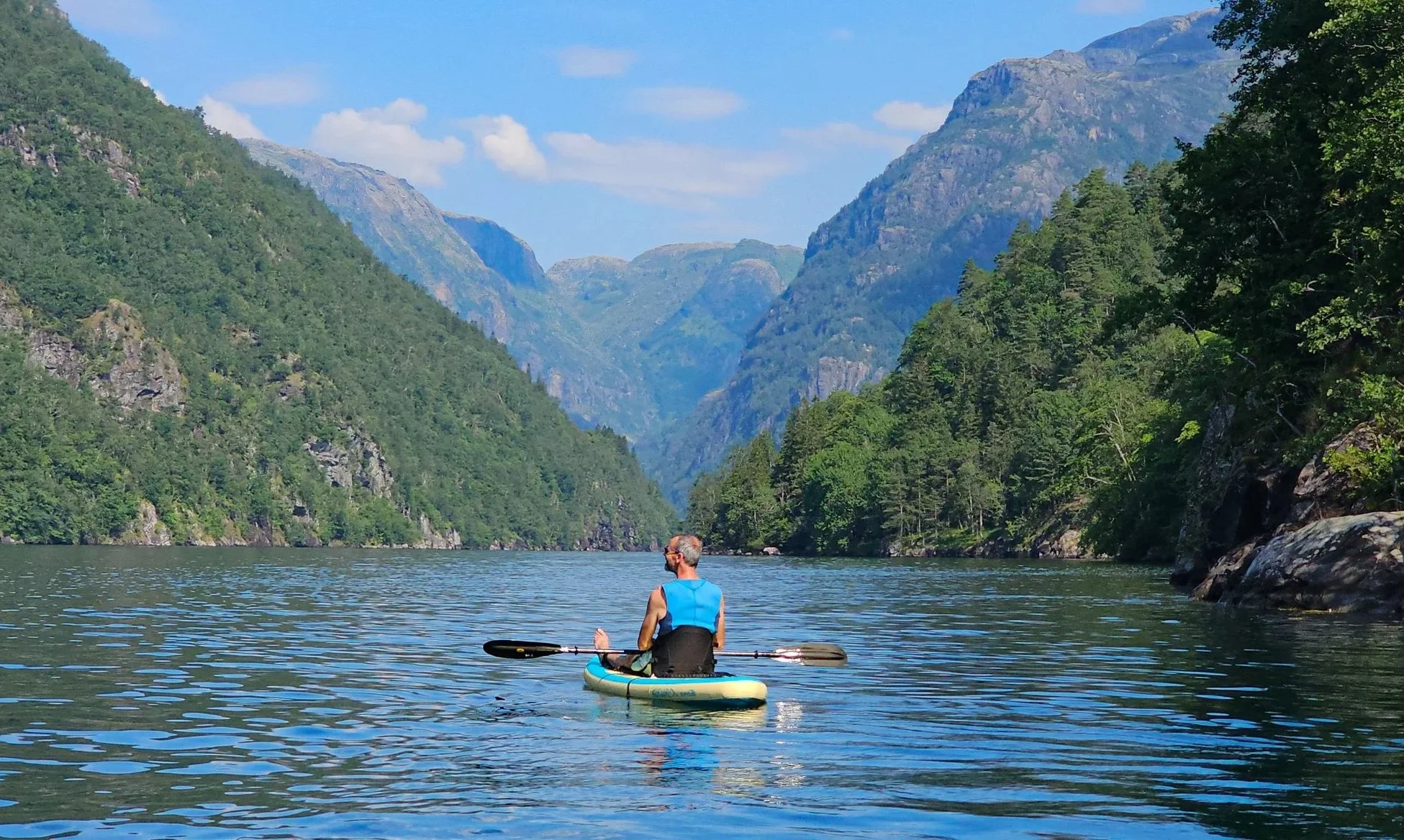 A man paddleboarding on a river surrounded by green mountains and a partly cloudy sky.