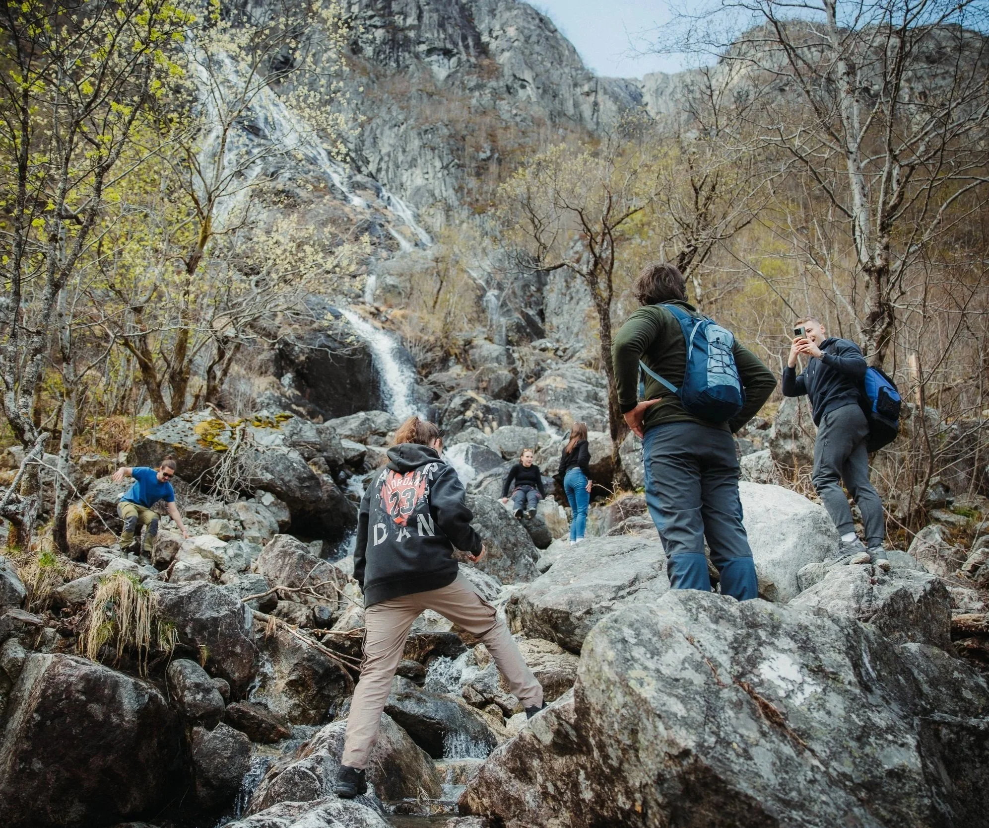 Group of people hiking and exploring a rocky area near a waterfall in a mountainous, wooded landscape during daytime.