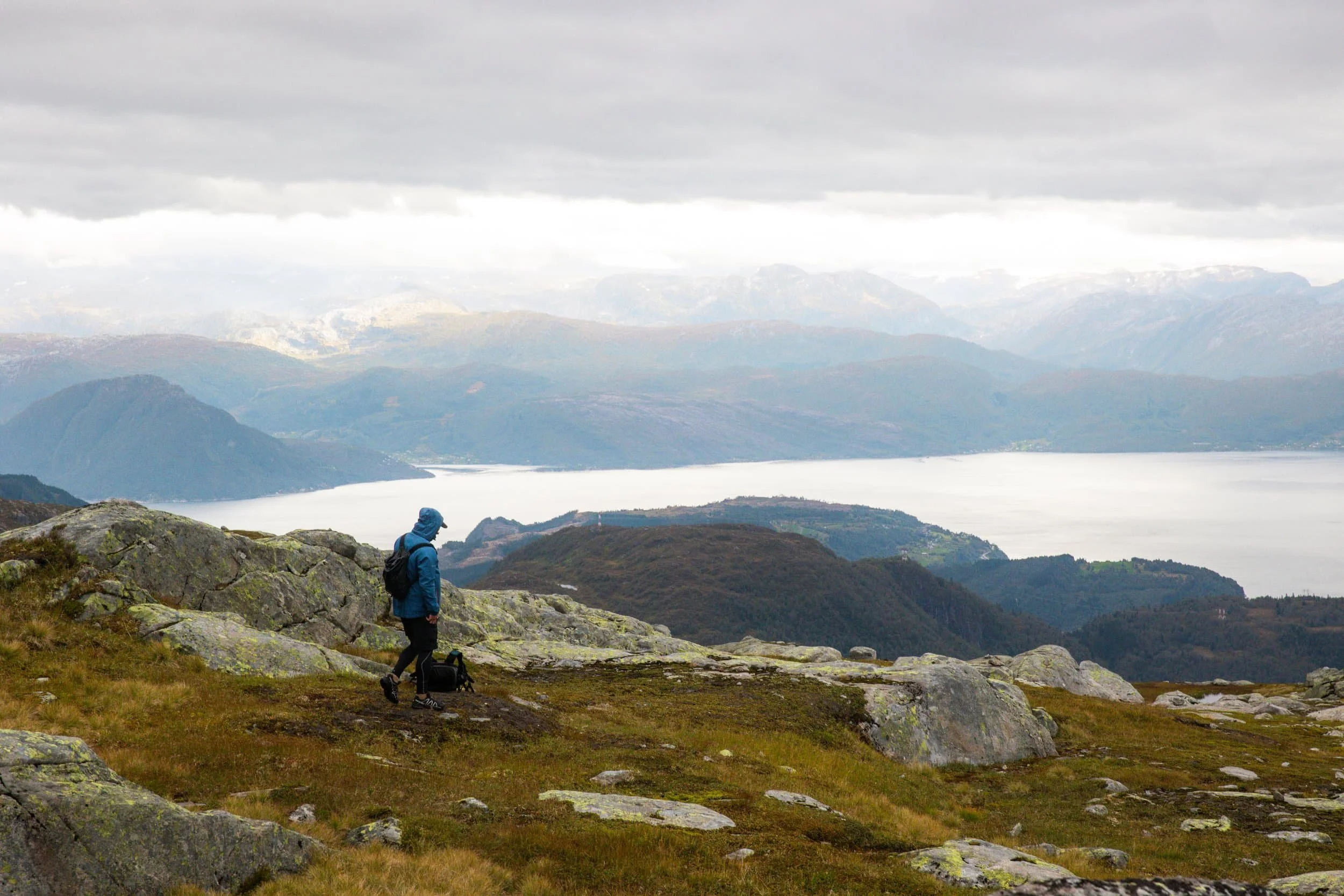 Hiker in blue jacket and backpack on rocky hillside overlooking a lake and mountains under cloudy sky.