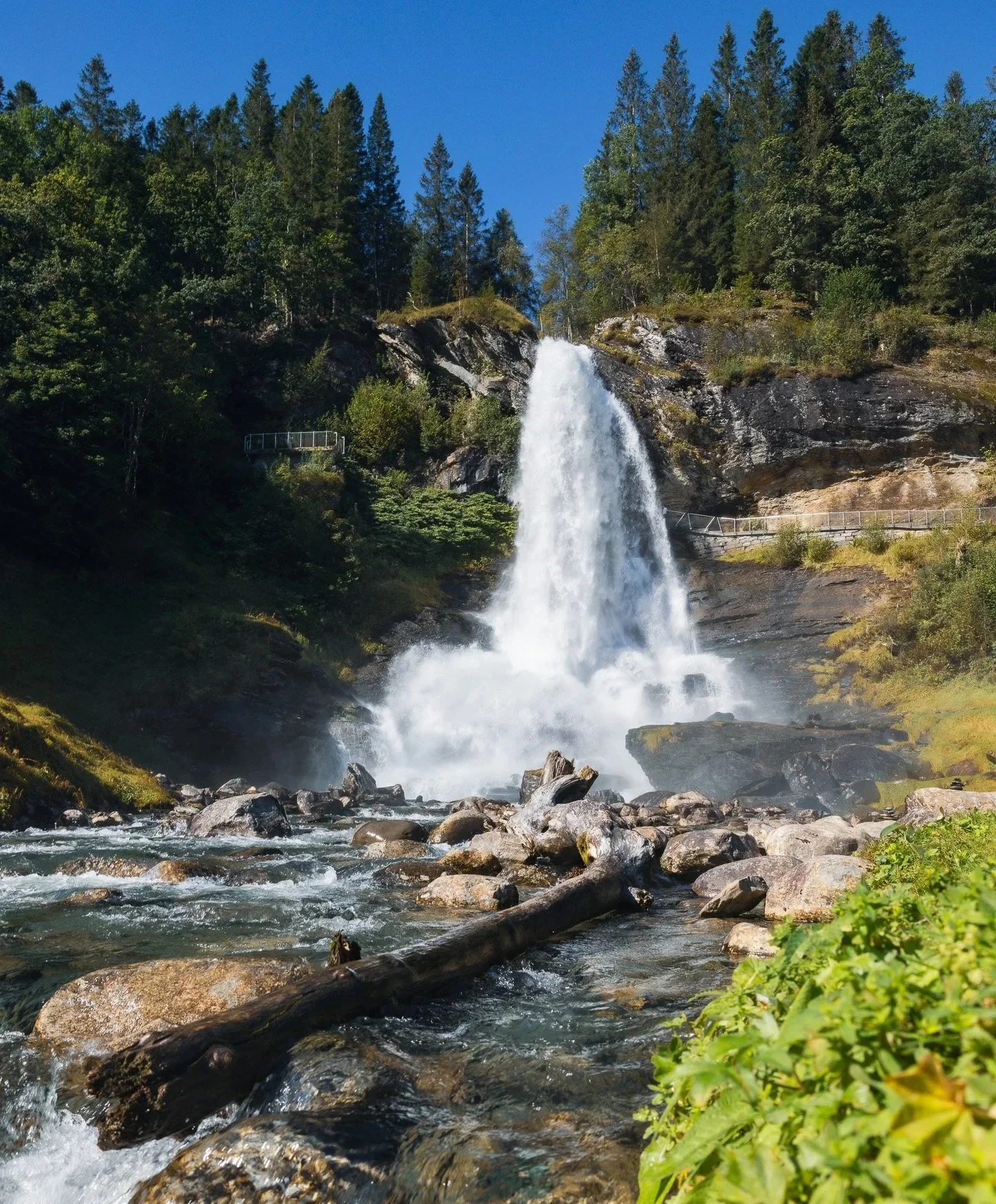 A tall waterfall cascading down a rocky cliff into a river below, surrounded by lush green trees on both sides, under a clear blue sky.