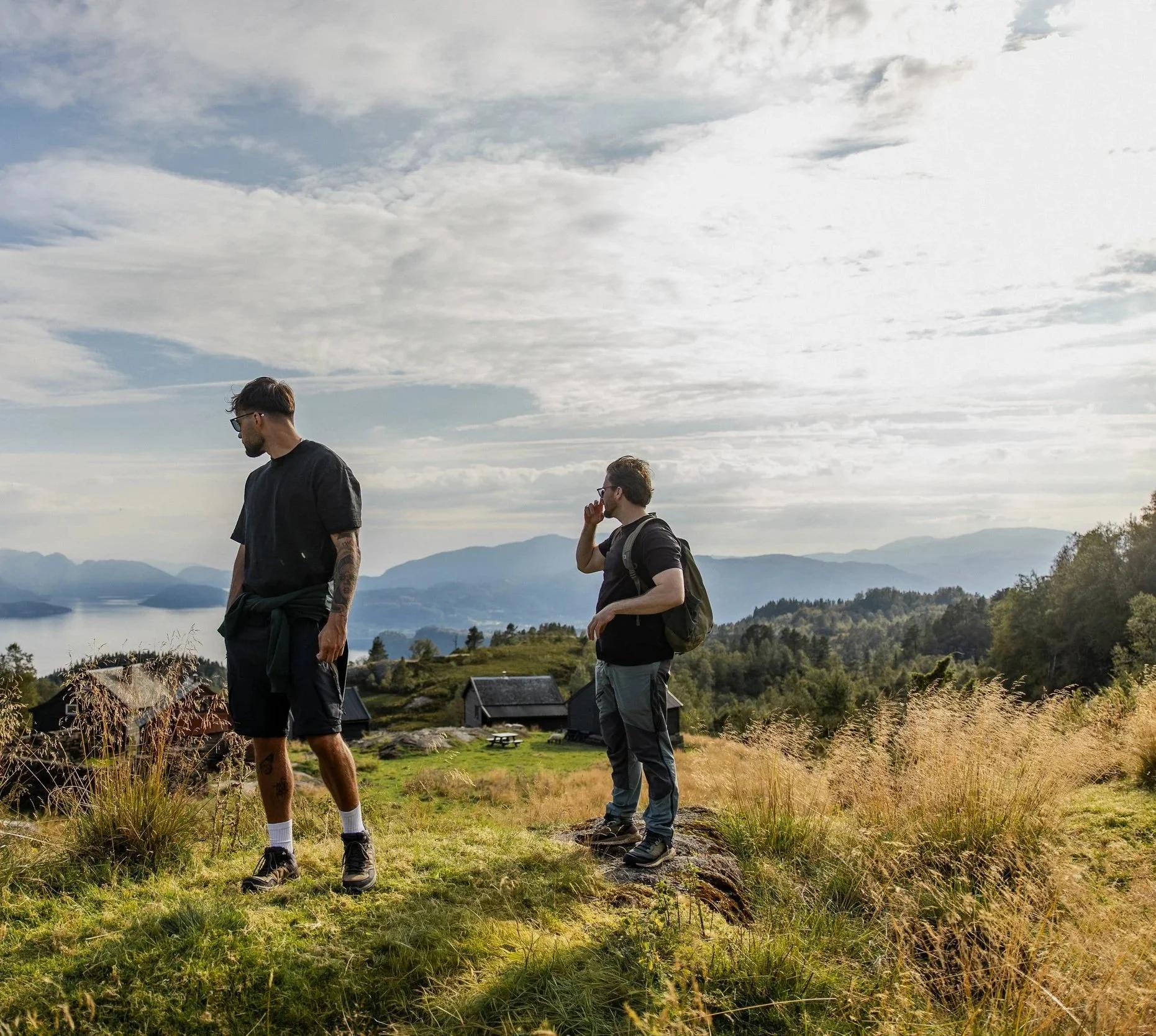 Two men are standing outdoors in a grassy area with hills, water, and houses in the background. One man has a beard and tattoos, wearing a black t-shirt and shorts, looking down. The other man is holding a phone to his ear, wearing glasses, a dark shirt, and jeans. The sky is partly cloudy.