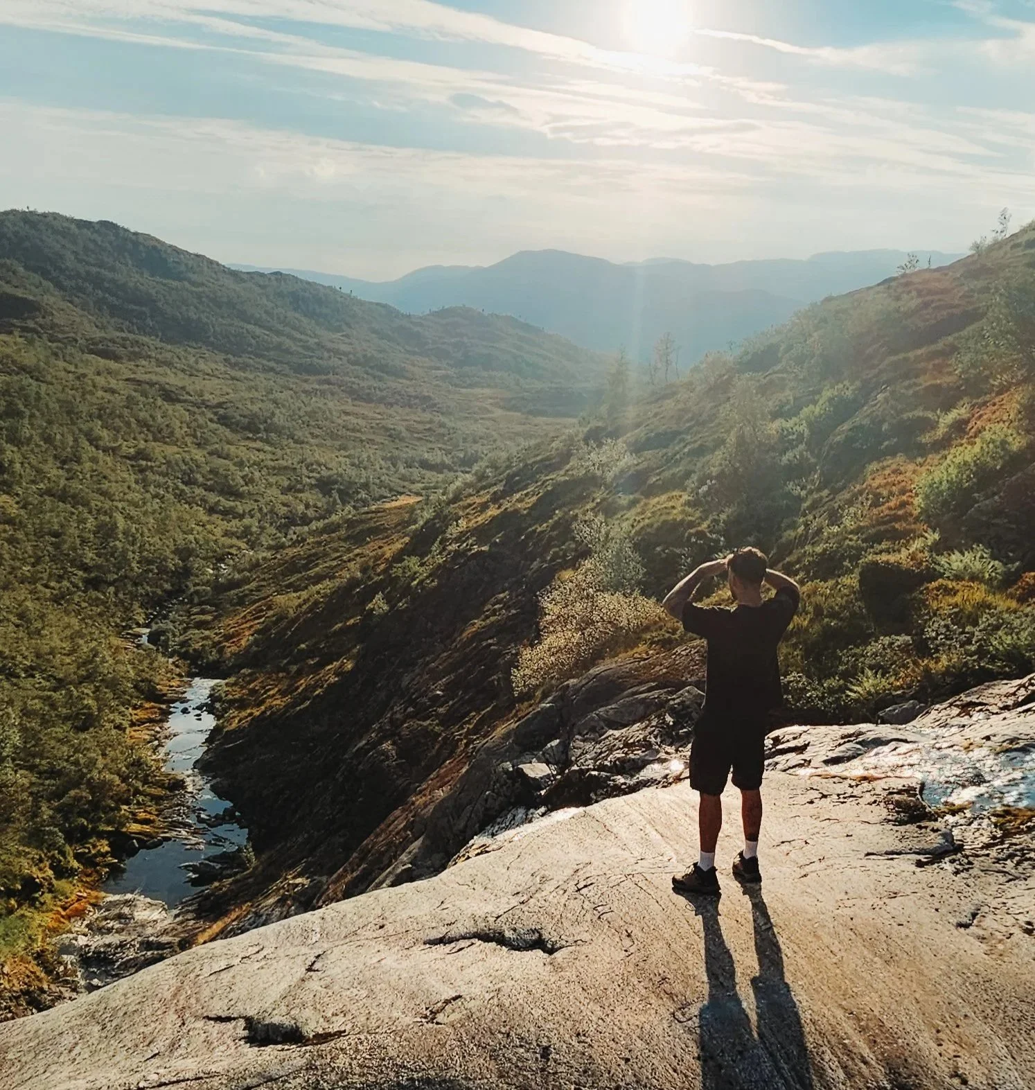 Person standing on a large rock overlooking a valley and mountains with a river flowing through it, under a partly cloudy sky.