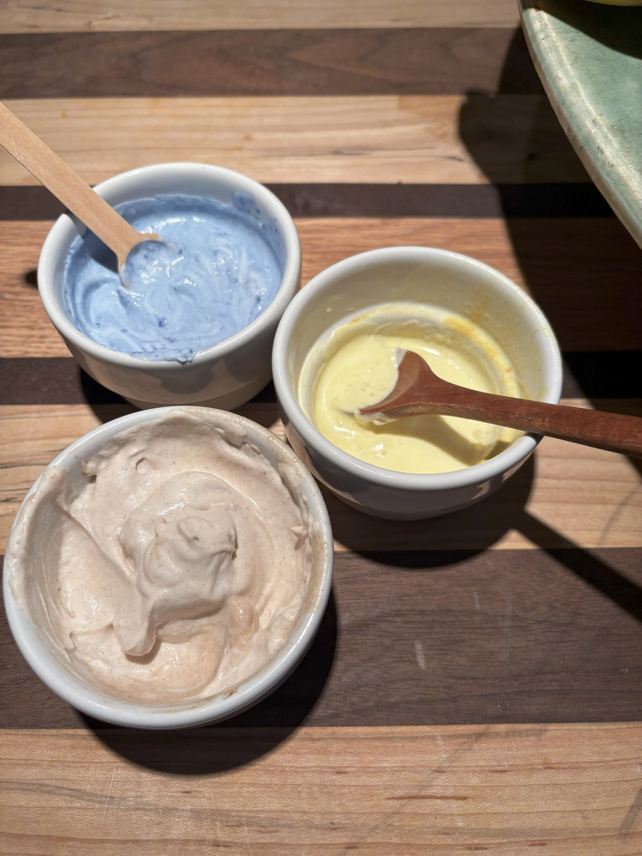 Three small bowls of different colored play dough on a wooden table, with spoons in each bowl.