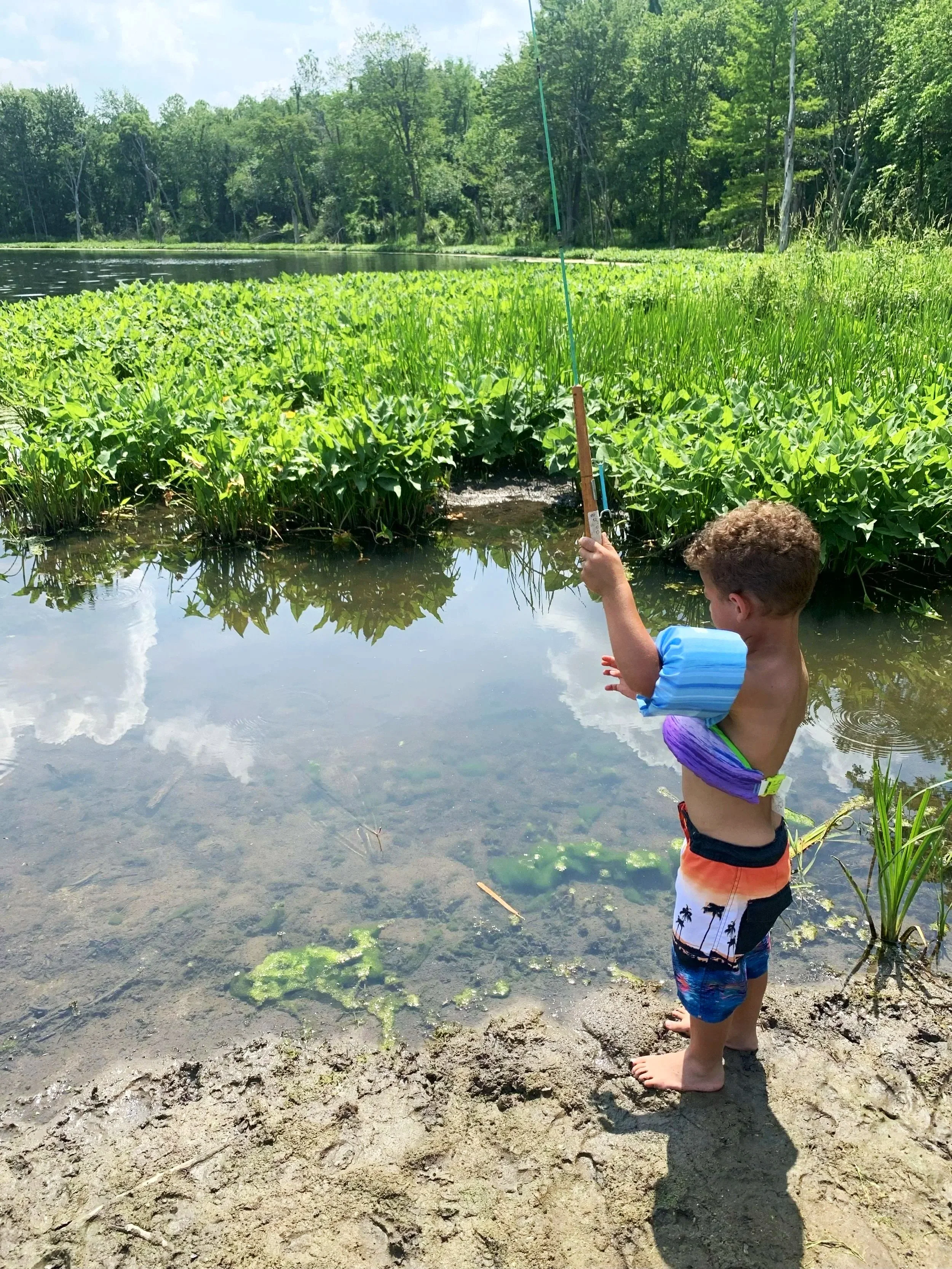 A young boy fishing by a lake on a sunny day, wearing swim trunks and a life jacket, with lush green trees and plants in the background.