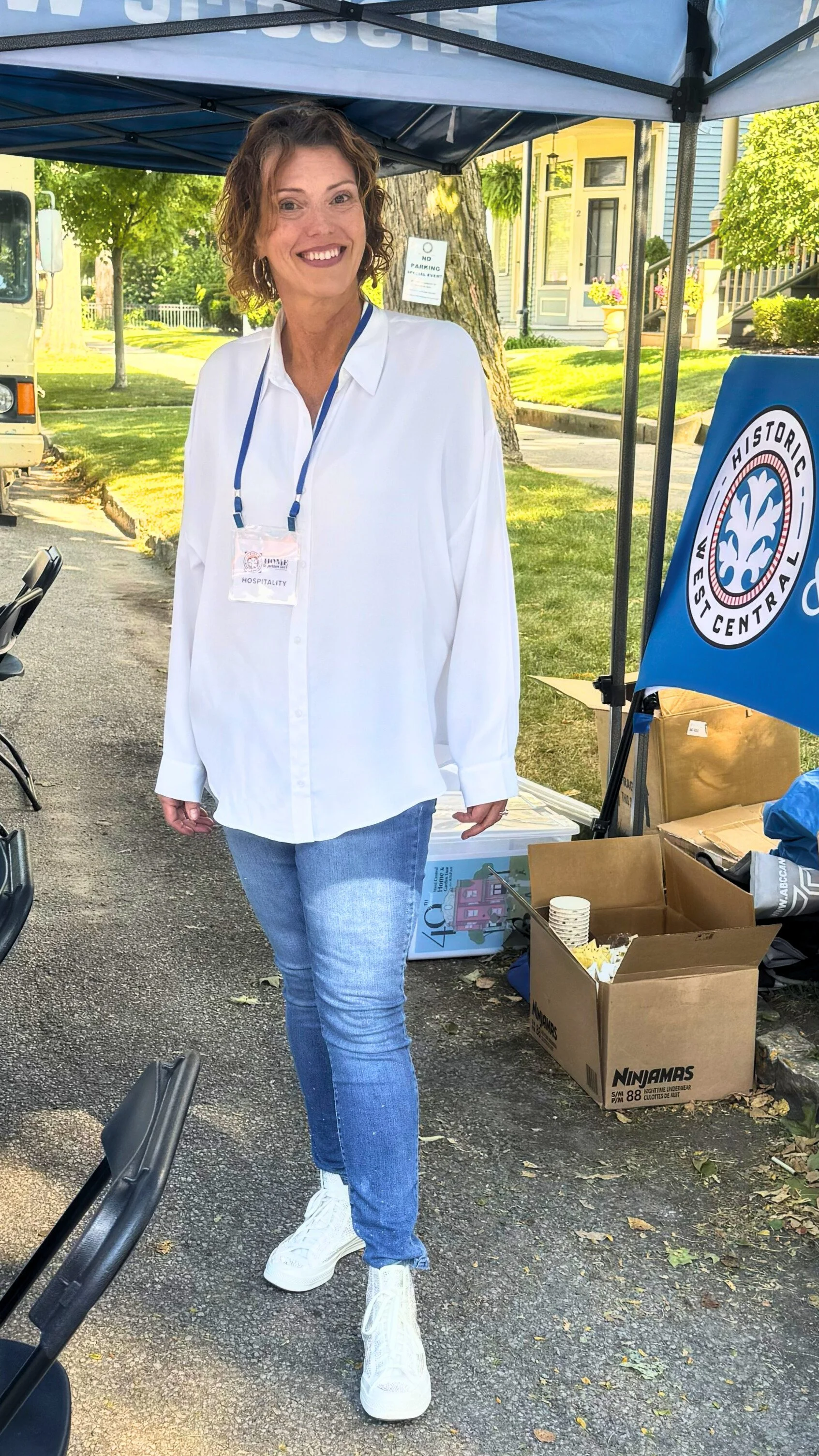 A woman in a white shirt and blue jeans standing outdoors under a blue canopy tent, smiling. There are boxes and a flag on a table nearby and trees and houses in the background.